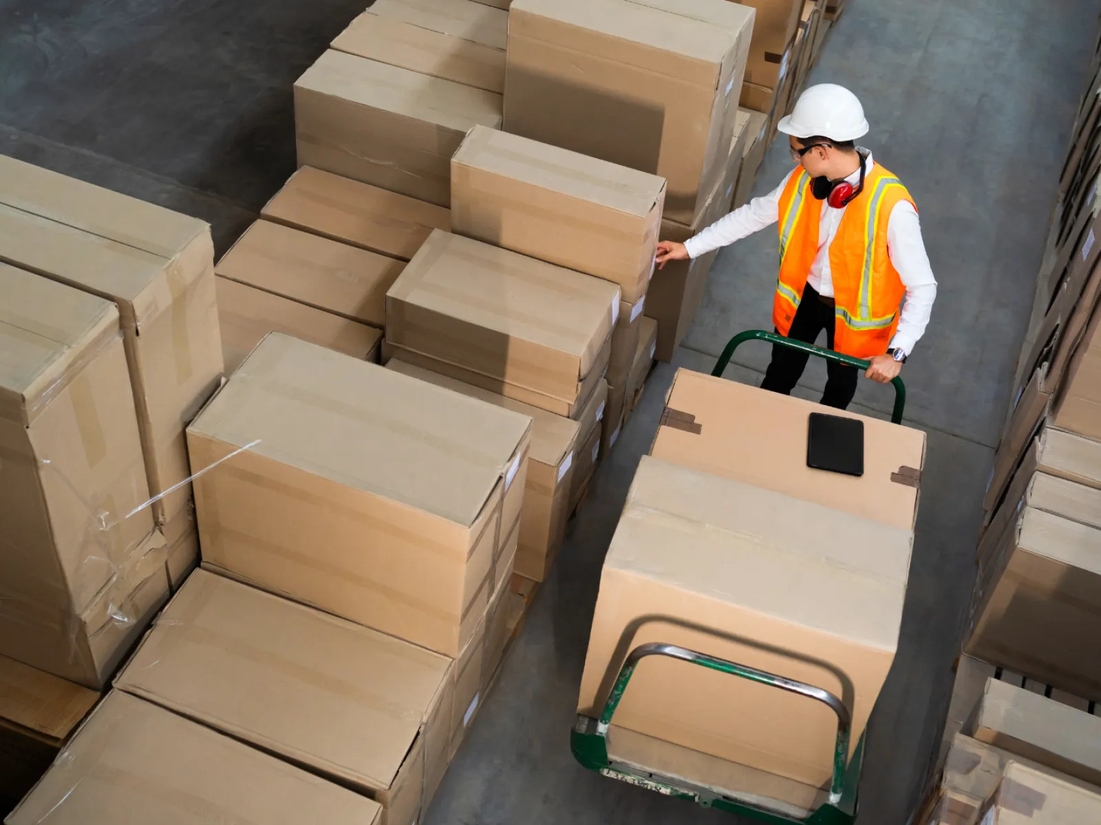 Warehouse worker in safety gear checking boxes on a hand truck surrounded by stacked cardboard packages.