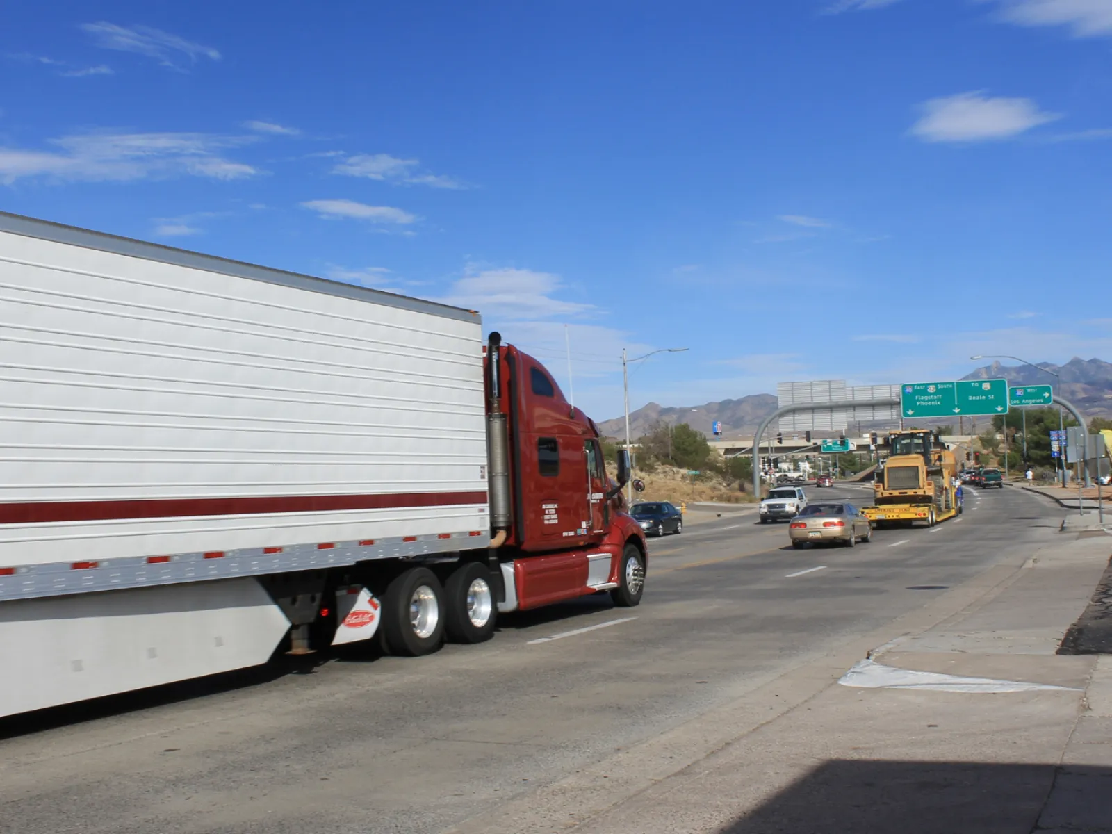 Red semi-truck with white trailer driving on a highway under a blue sky with mountains in the background
