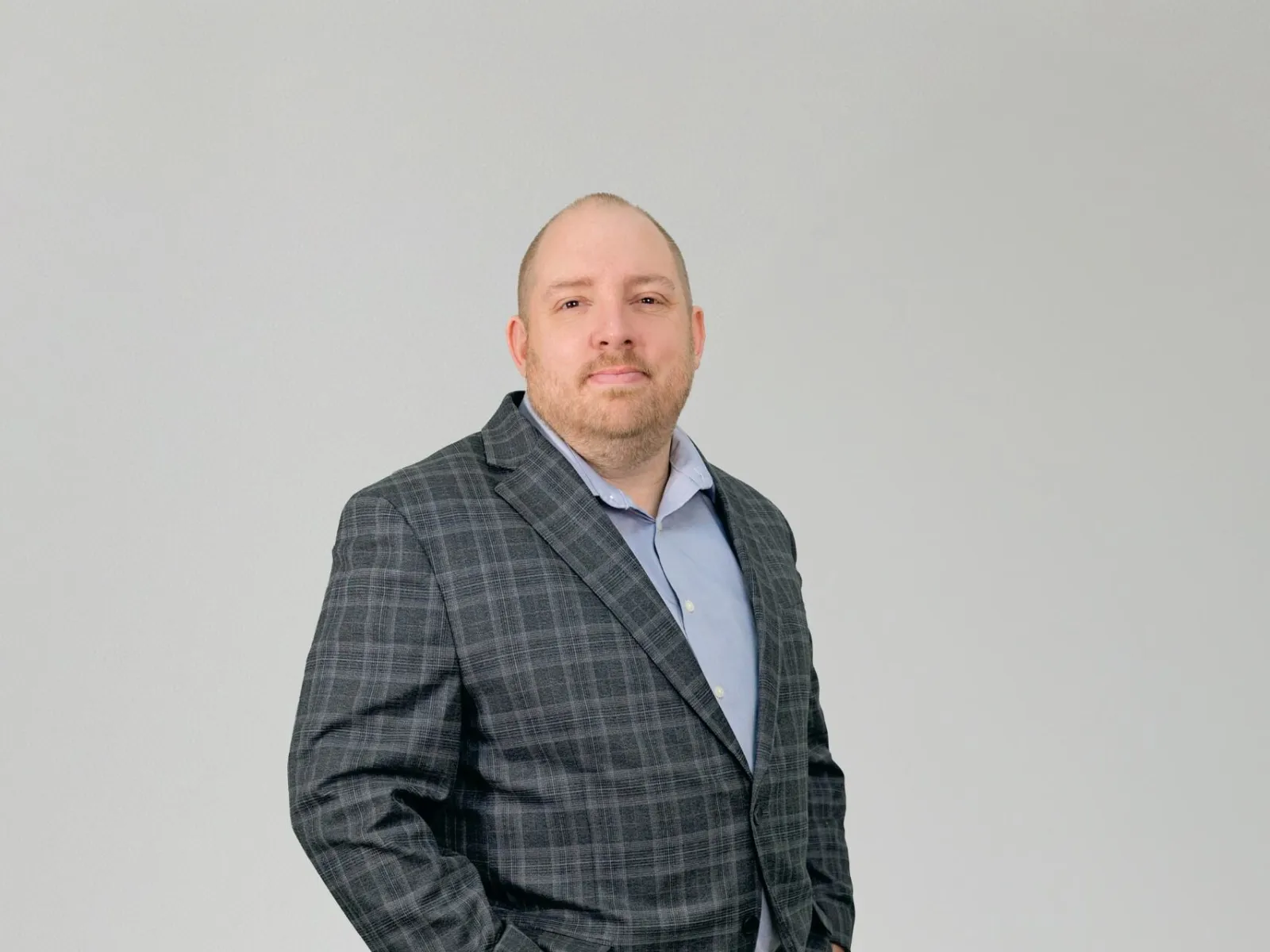Man wearing a gray plaid blazer, light blue shirt, jeans, and black shoes standing with hands in pockets