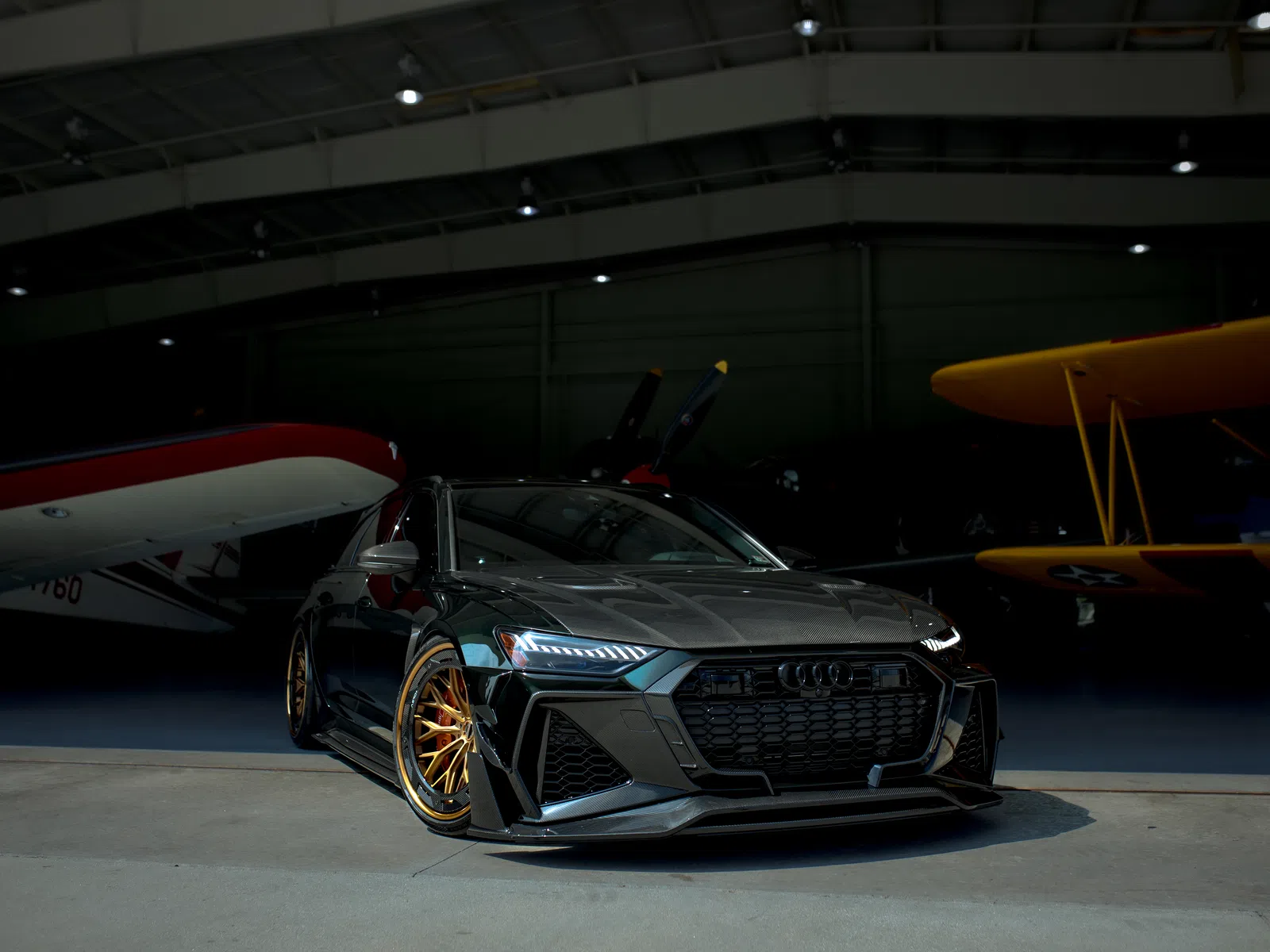 Sleek black Audi sports car with gold rims parked in an aircraft hangar beside vintage planes.