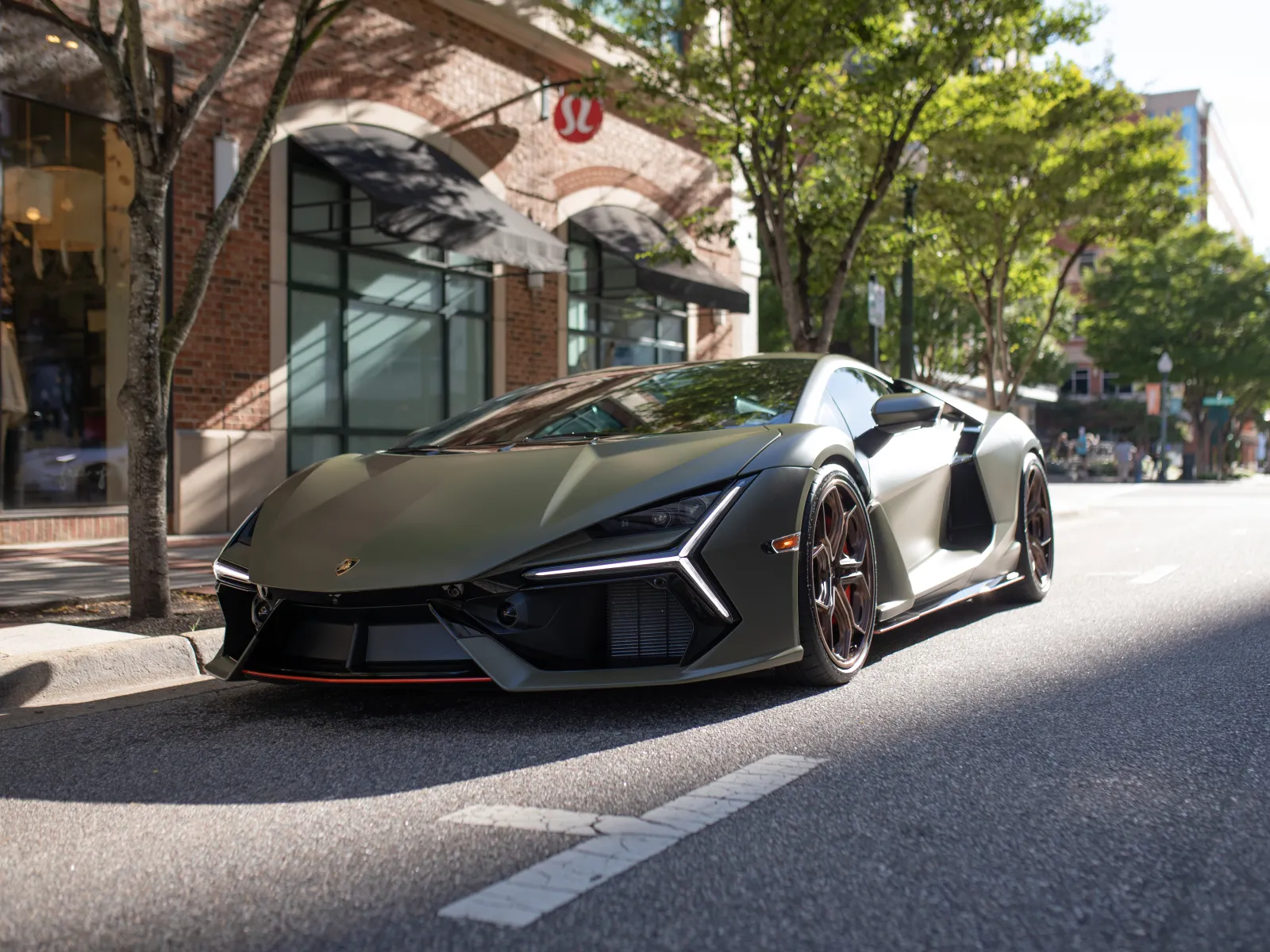 Matte green Lamborghini sports car parked on a sunny urban street near brick buildings and trees.