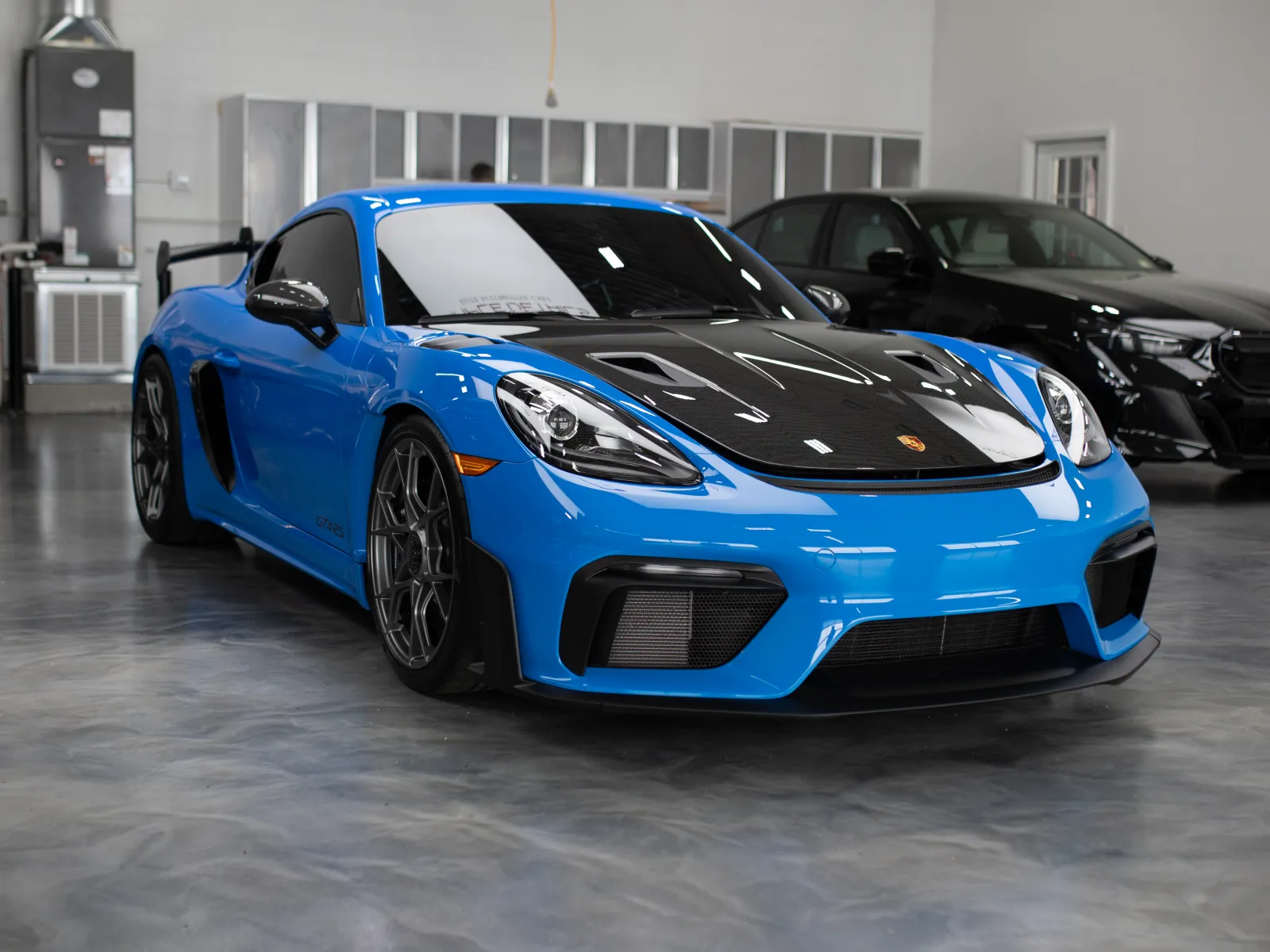 Blue Porsche sports car with black hood parked in a clean, well-lit indoor showroom with polished concrete floor.