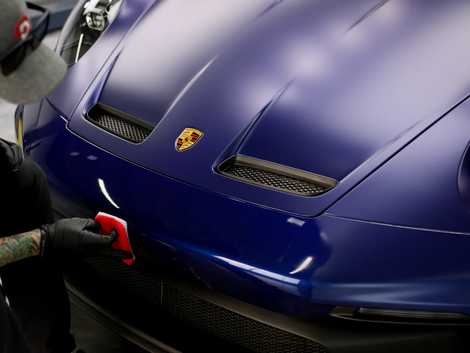 Man polishing the front hood of a blue Porsche sports car in a garage with a red cloth.