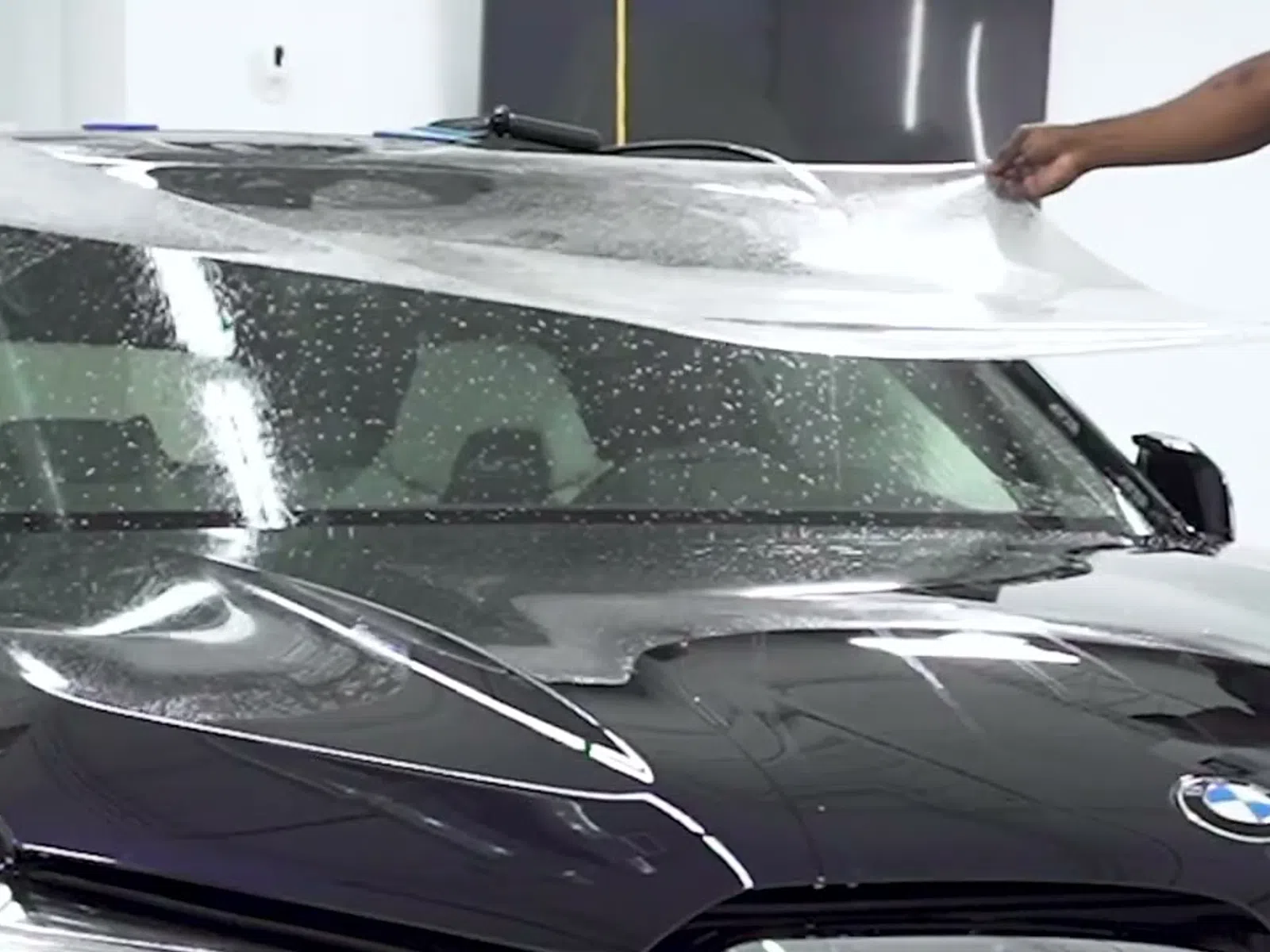 Person applying clear protective film to black BMW car hood and windshield inside a garage or workshop.