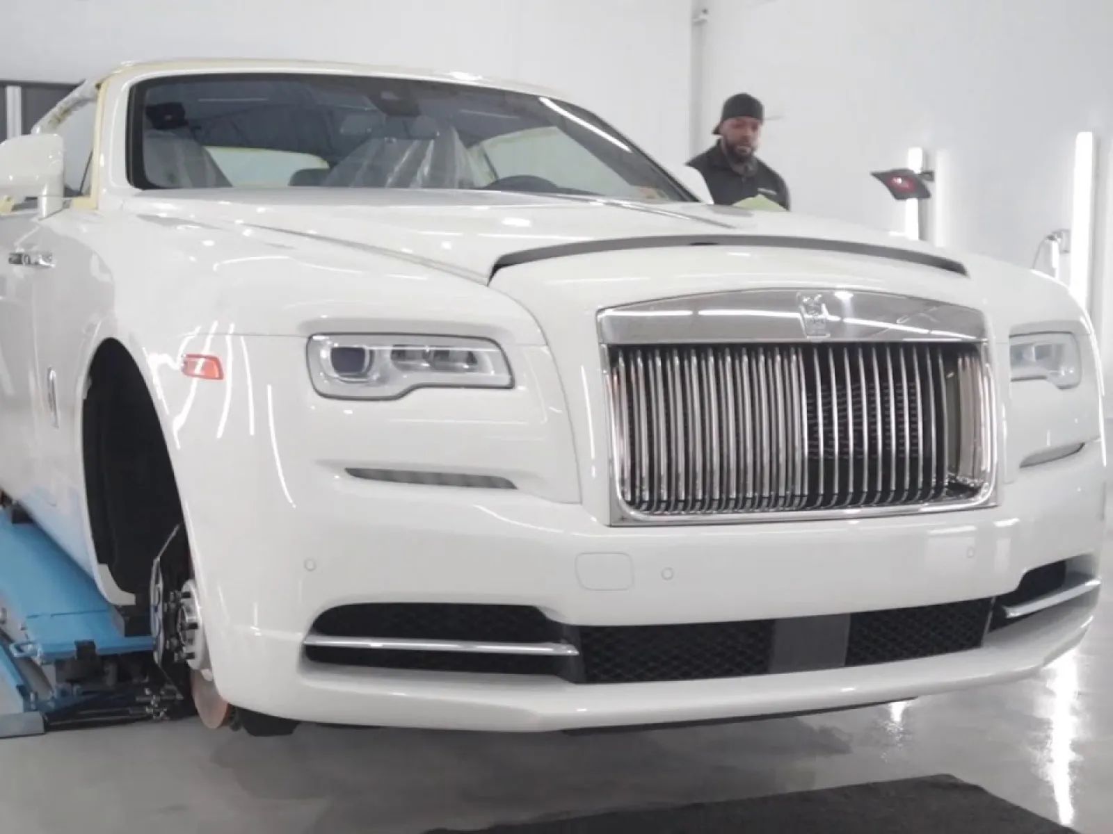 White luxury Rolls-Royce car lifted in a bright auto workshop with a mechanic working in the background.