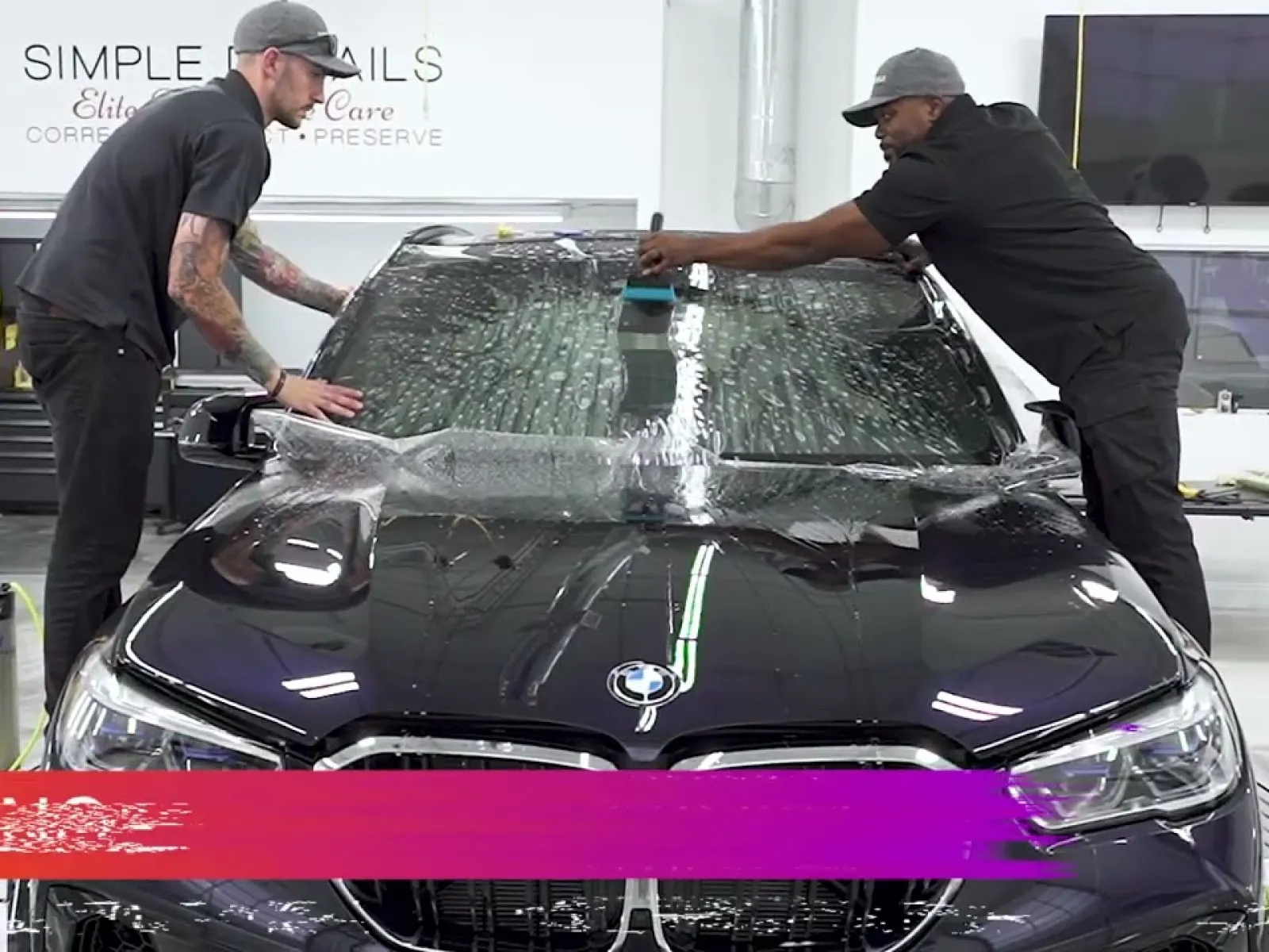 Two men applying protective film to the windshield of a black BMW in a clean auto care workshop.