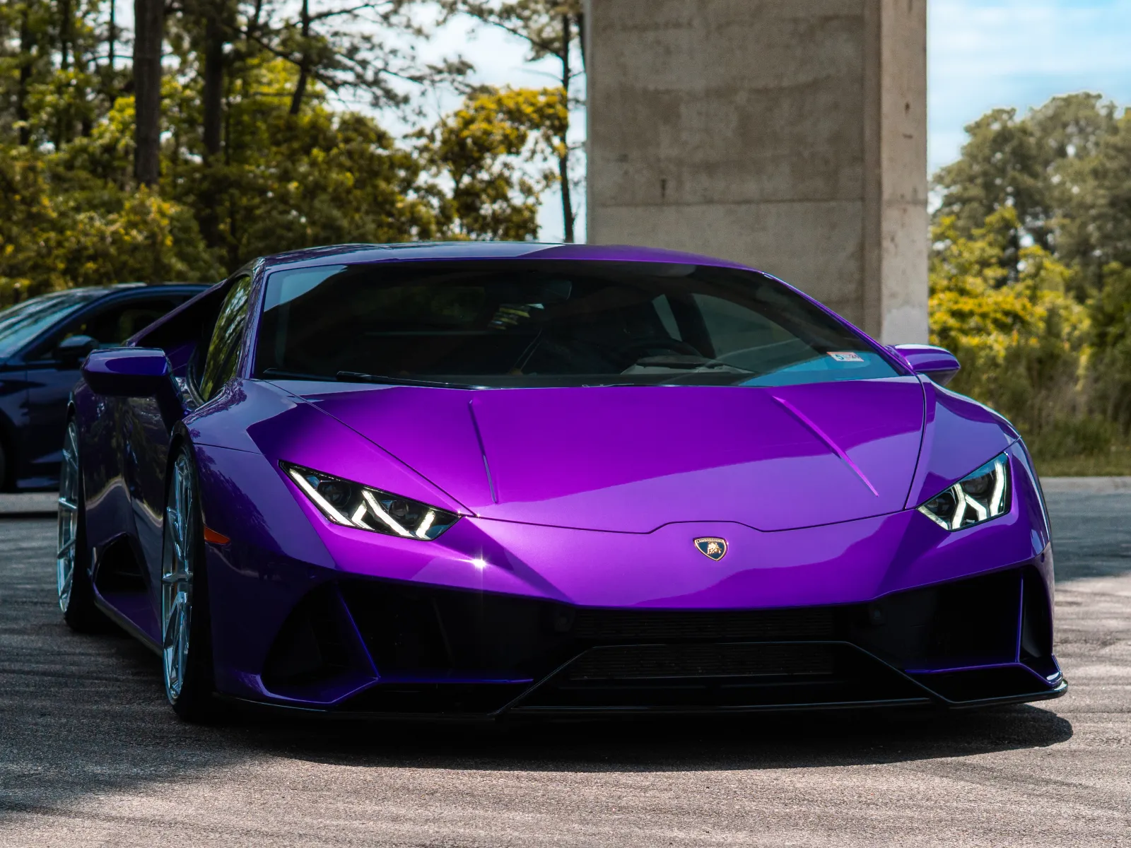 Purple Lamborghini sports car parked on road with black car and trees in background under blue sky.