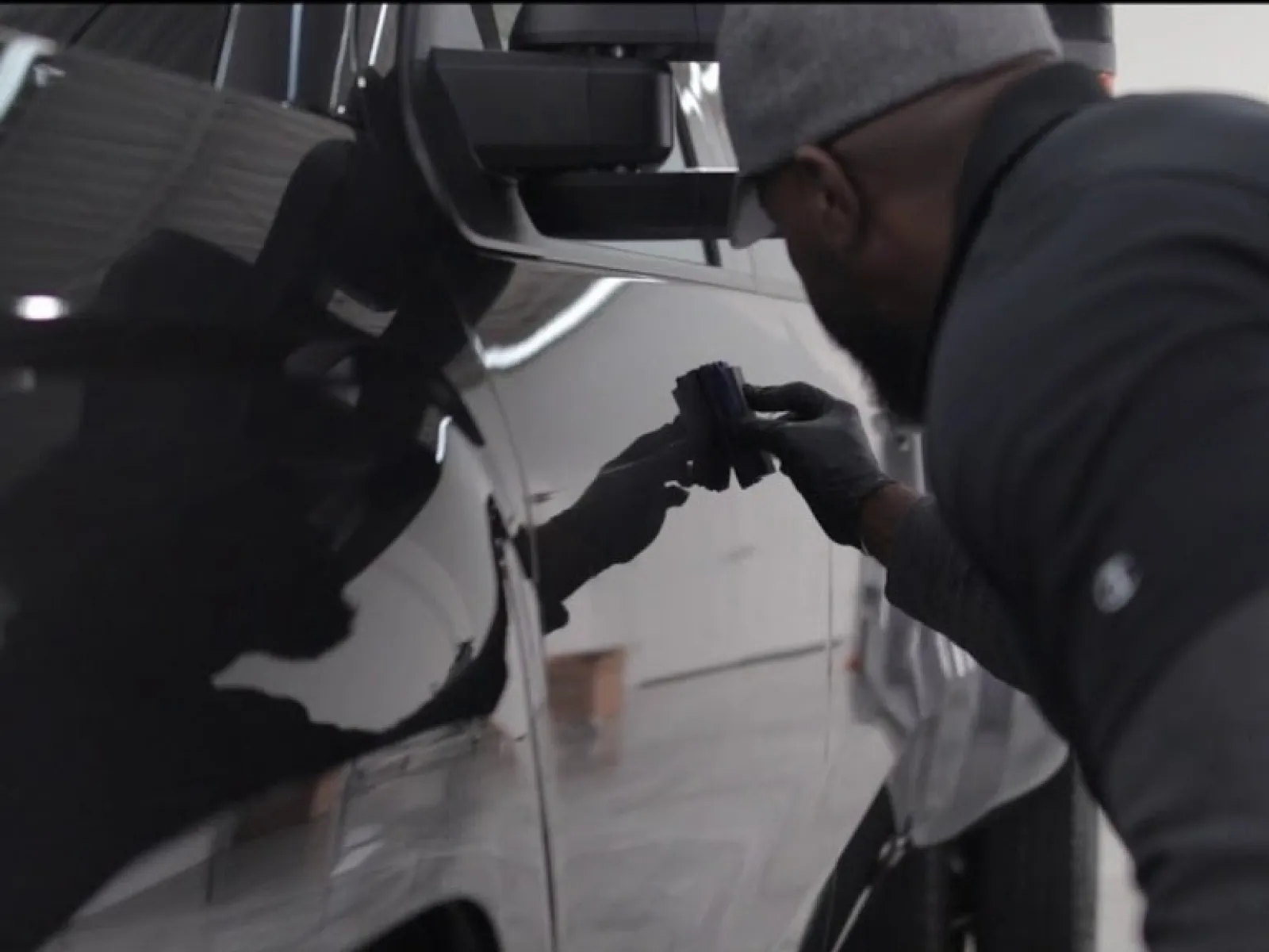 Man wearing gloves inspects the side panel of a black car in a well-lit garage.
