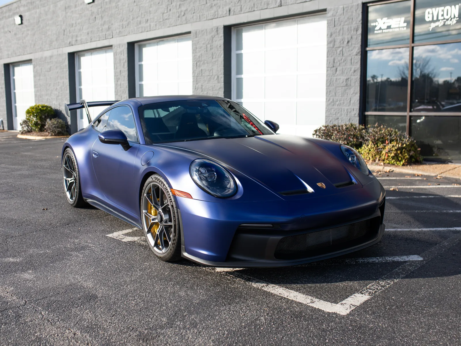 Matte blue Porsche sports car parked outside a modern garage on a sunny day.