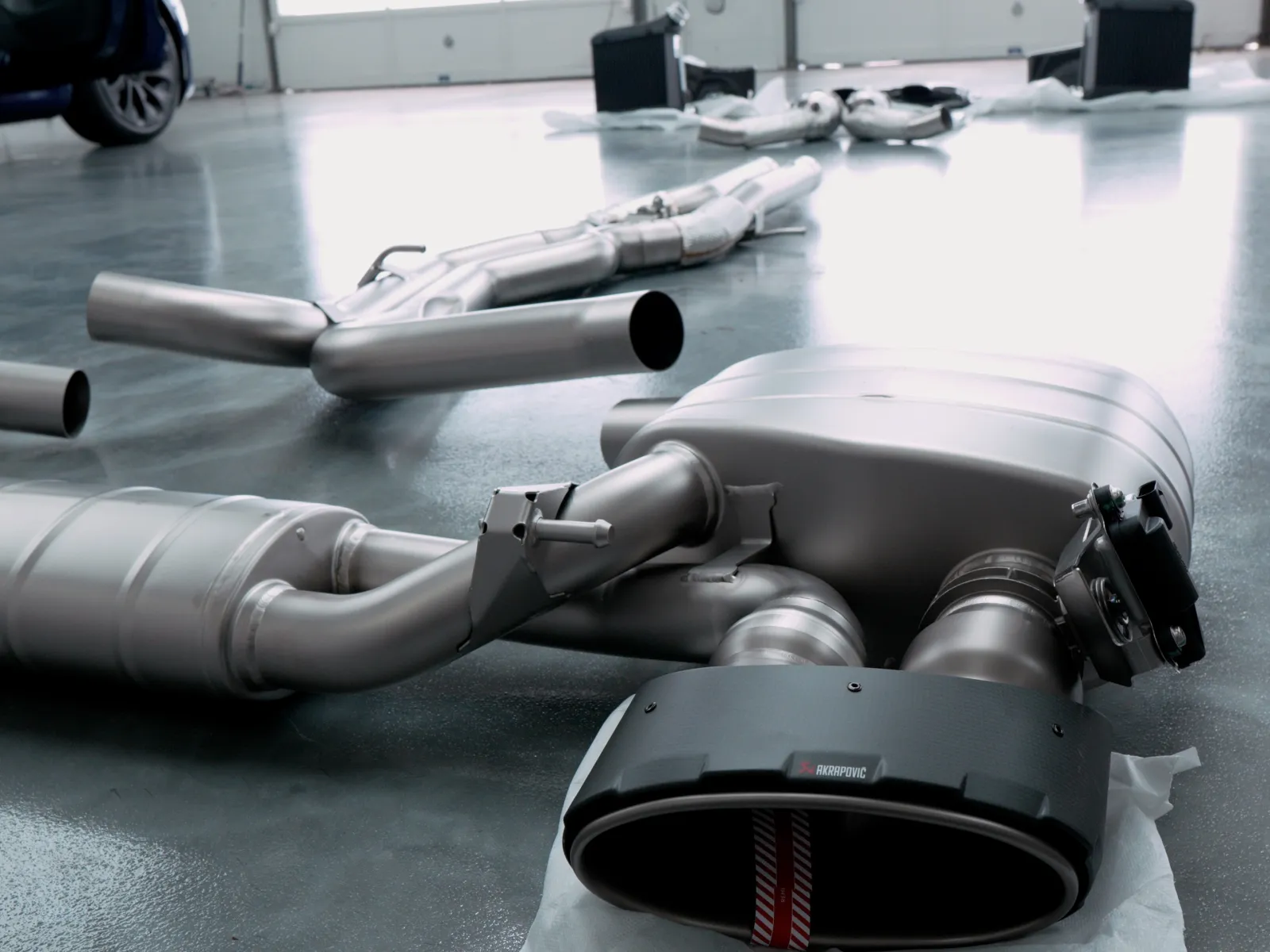 Car exhaust system components including mufflers and pipes laid out on a workshop floor with a car nearby.