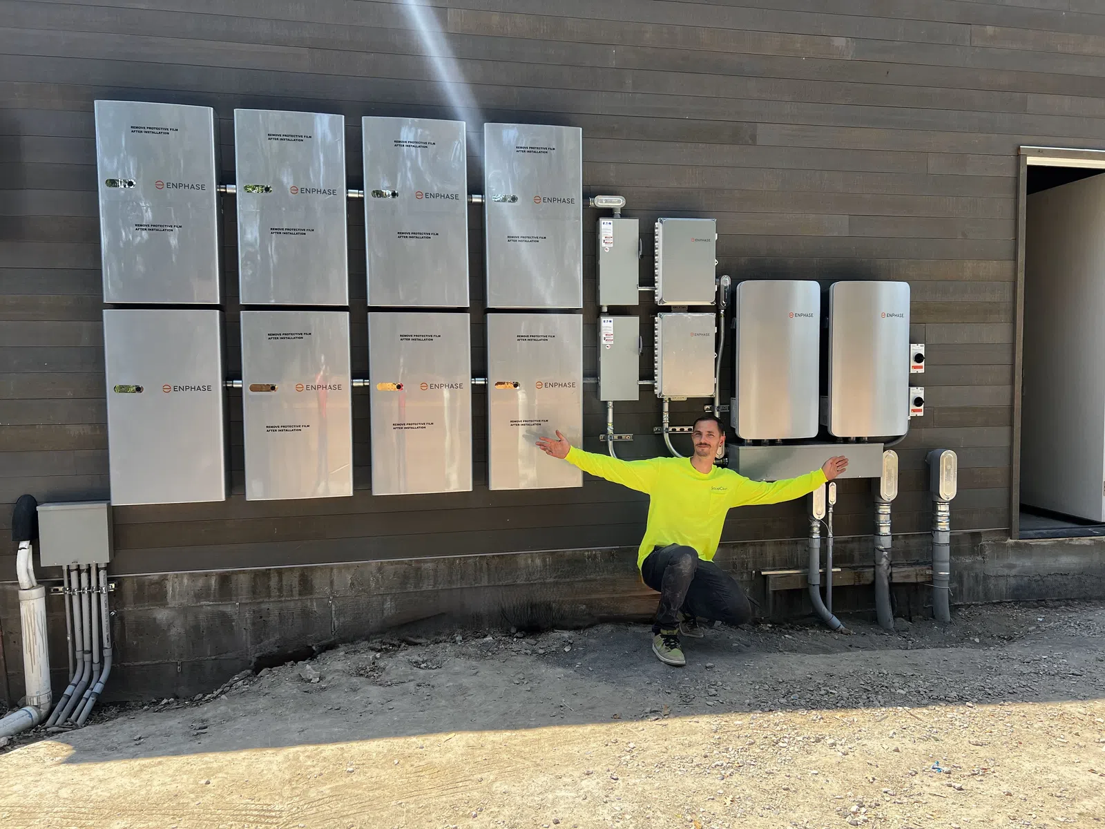 Man in neon yellow shirt kneeling by multiple Enphase electrical panels mounted on a brown exterior wall outdoors.