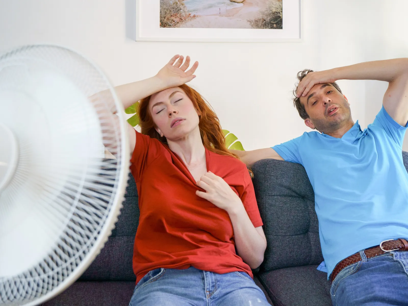 Man and woman sitting on couch feeling hot and exhausted with fan in foreground during heatwave indoors.