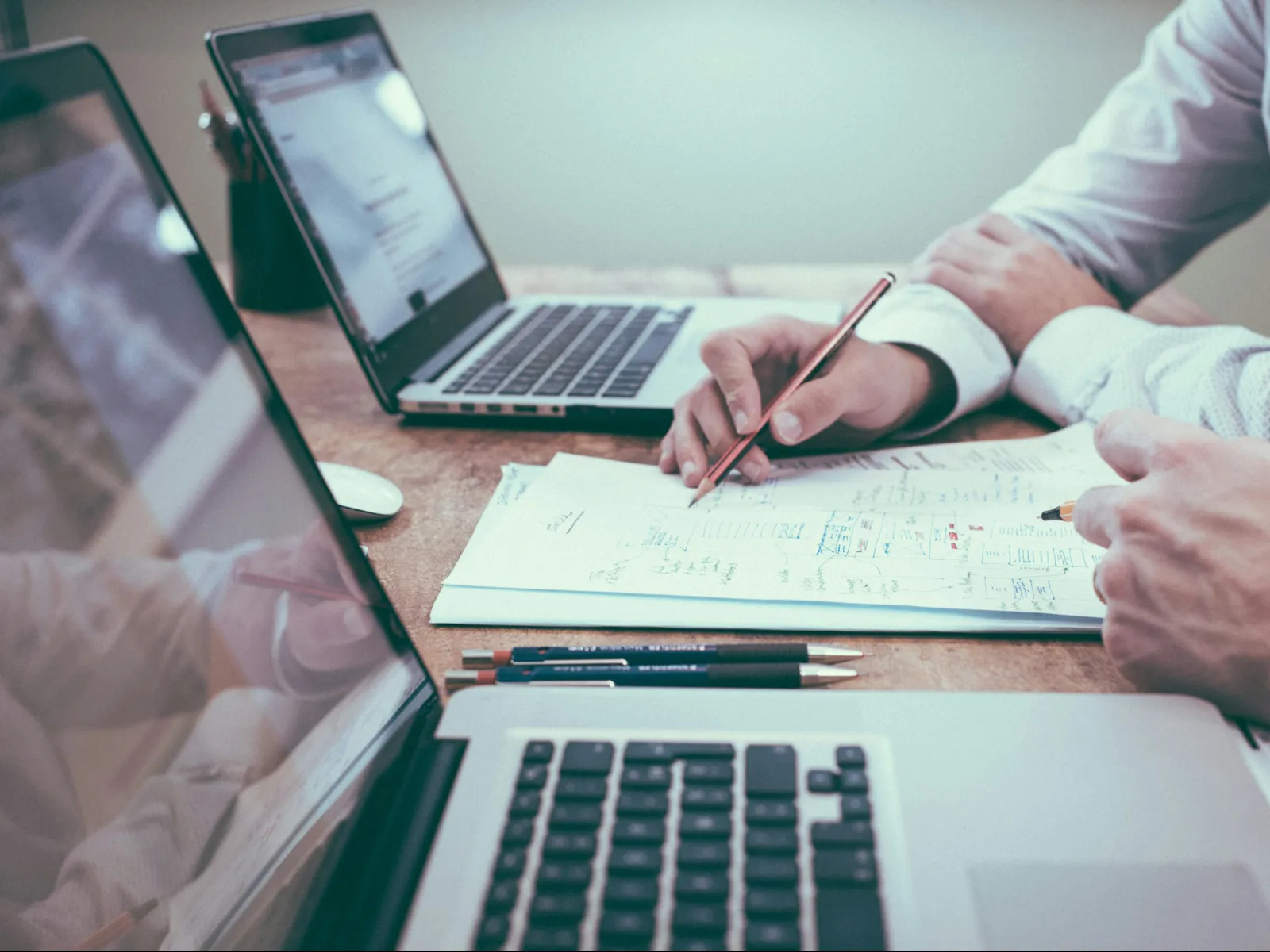 Two people collaborating on project plans with laptops and paperwork on a wooden desk.