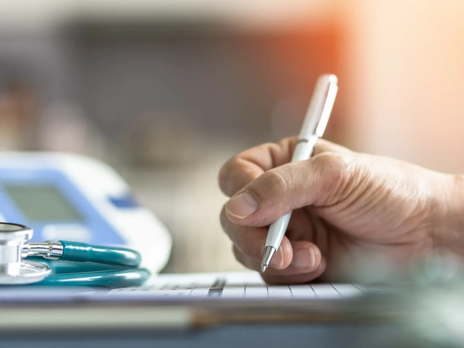 Close-up of a hand writing on paper with a stethoscope and blood pressure monitor in the background