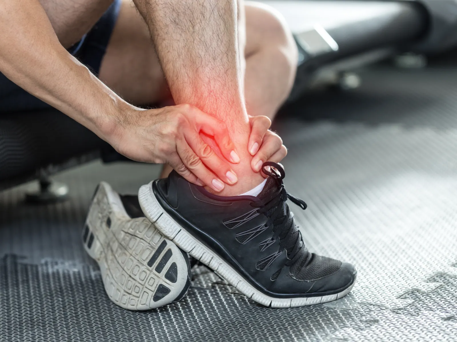 Person in black sneakers holding their painful ankle with red pain highlight on gym floor mat.