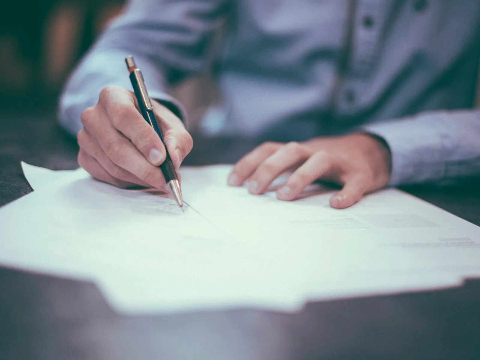 Person writing on a document with a pen on a desk, wearing a light blue long-sleeve shirt.