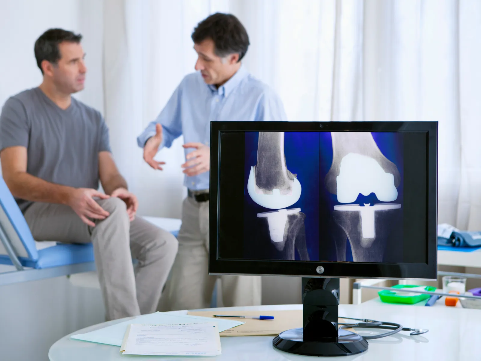 Doctor consulting male patient with knee pain and showing knee X-ray on monitor in medical clinic.