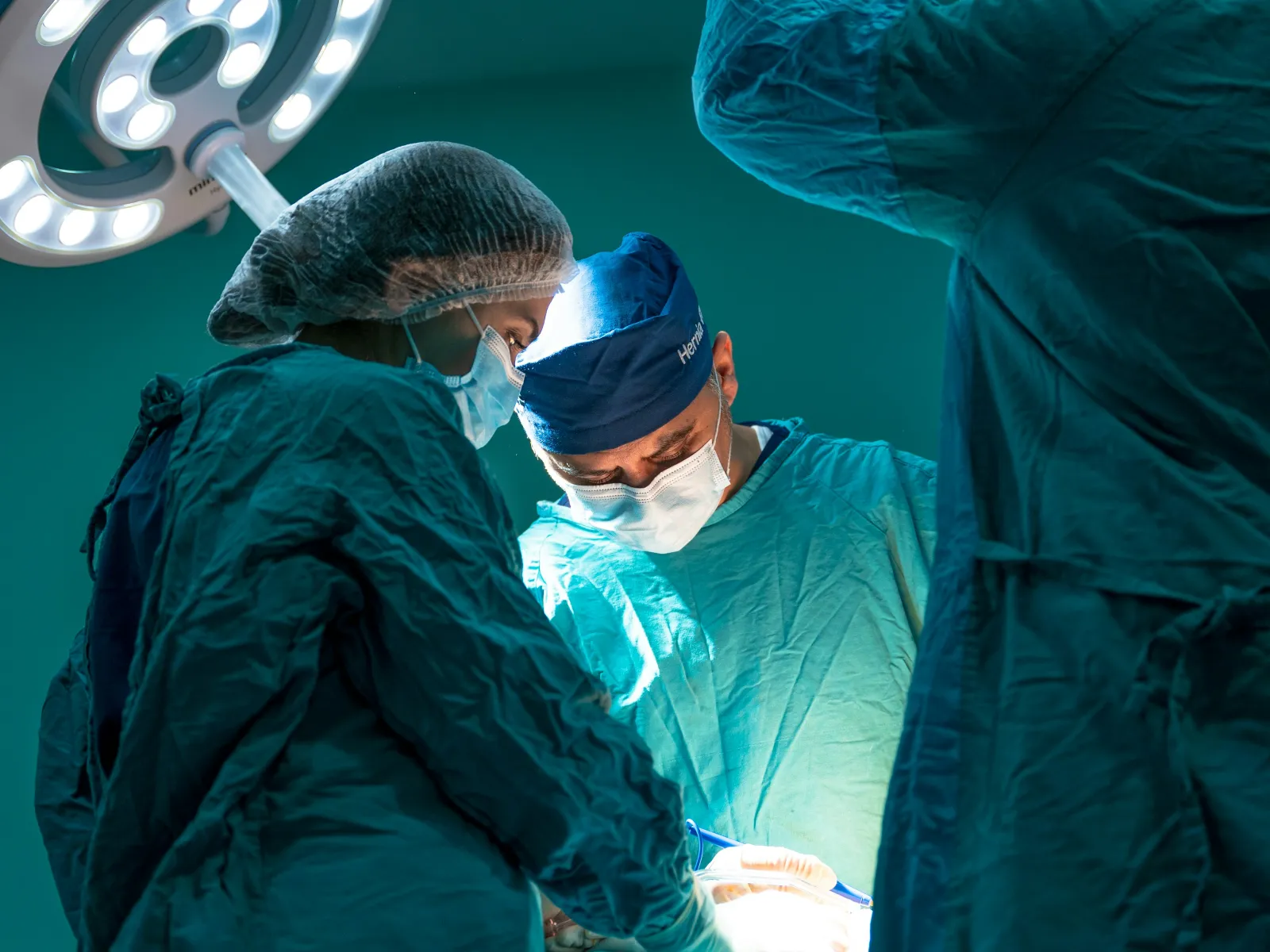 Surgeons in blue scrubs perform surgery under bright operating room lights in a sterile environment