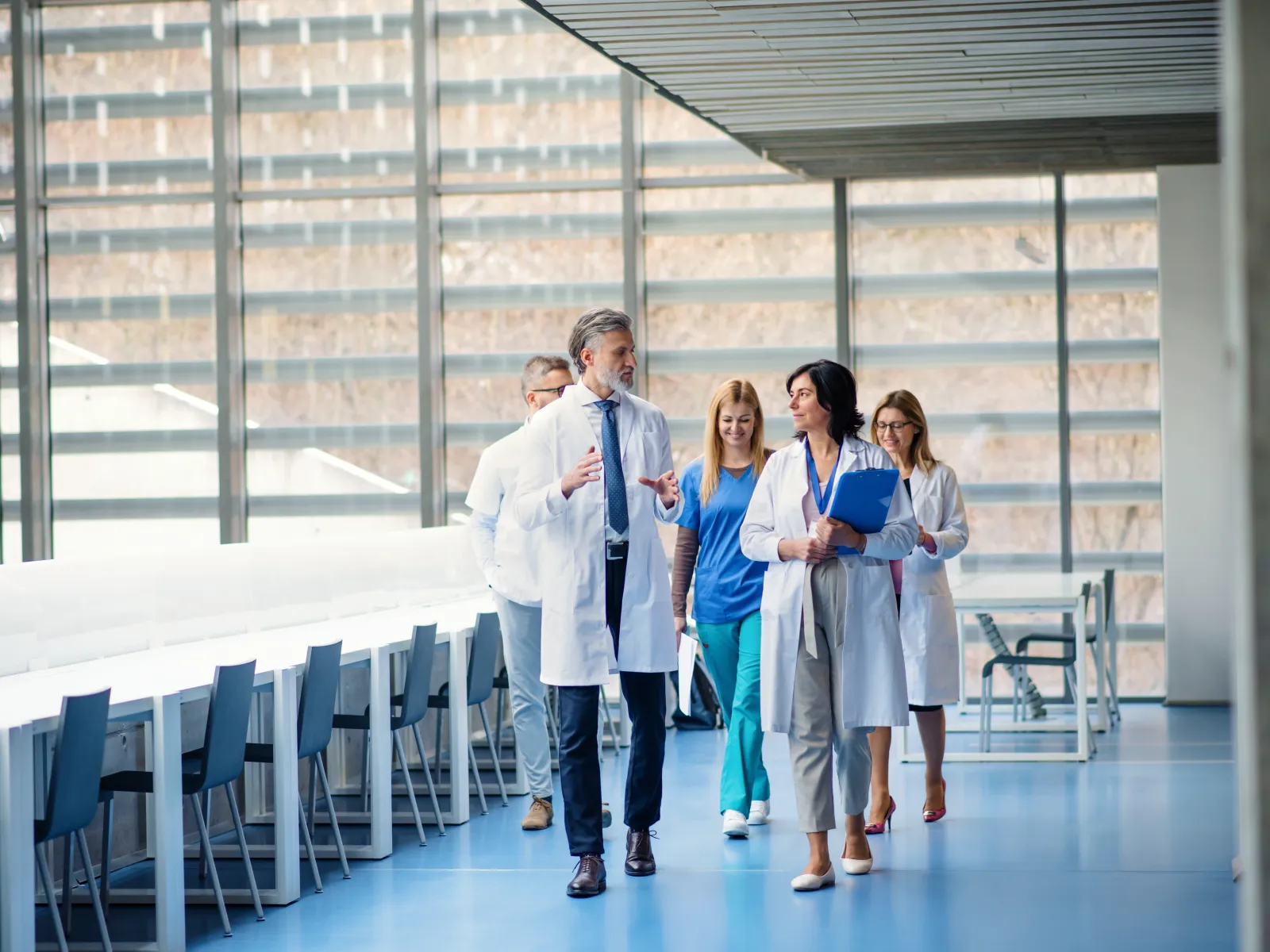 Group of doctors and nurses walking and discussing in a bright modern hospital corridor with large windows.