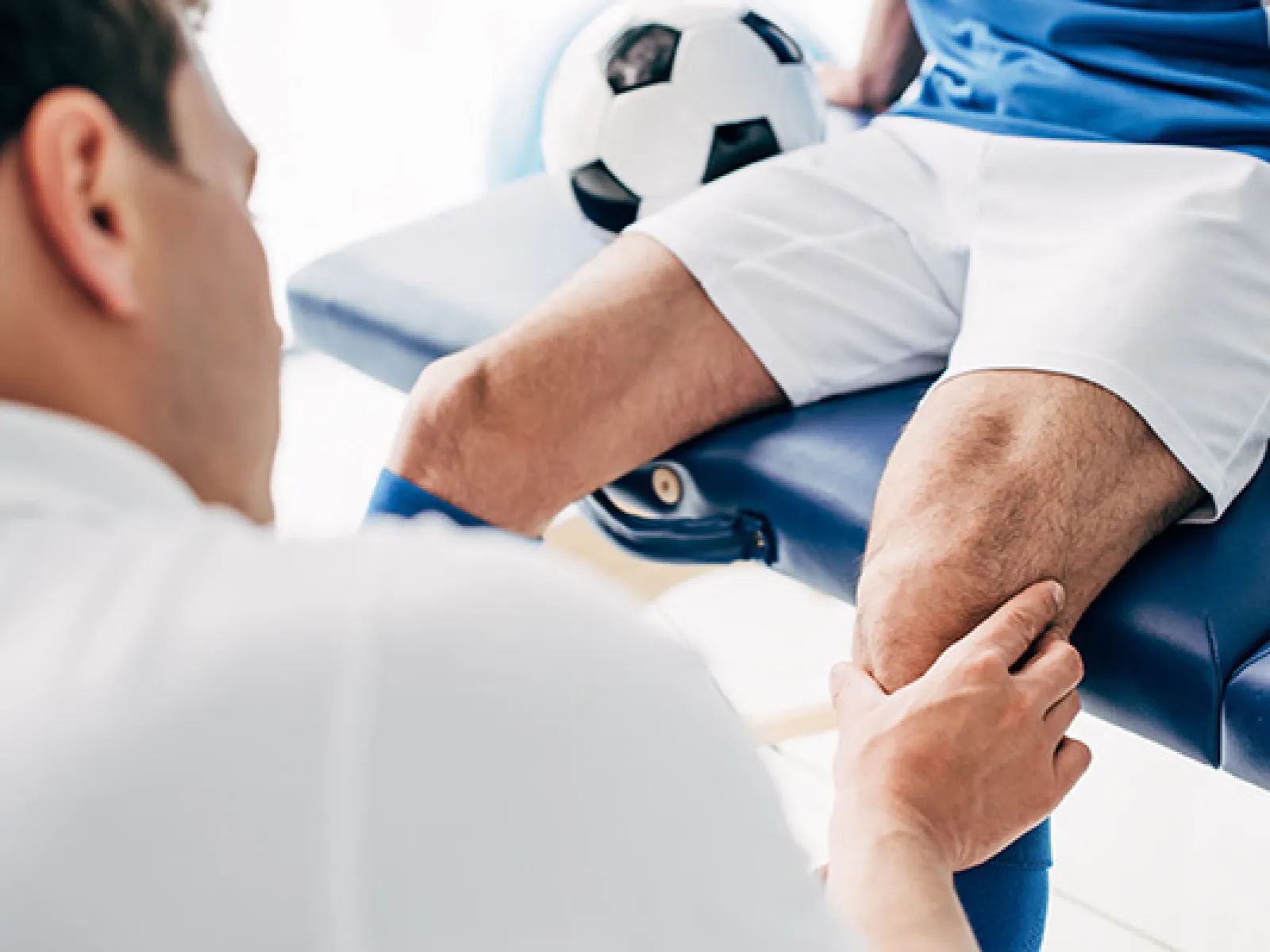 Physiotherapist examining soccer player's knee with a ball nearby on treatment table indoors.