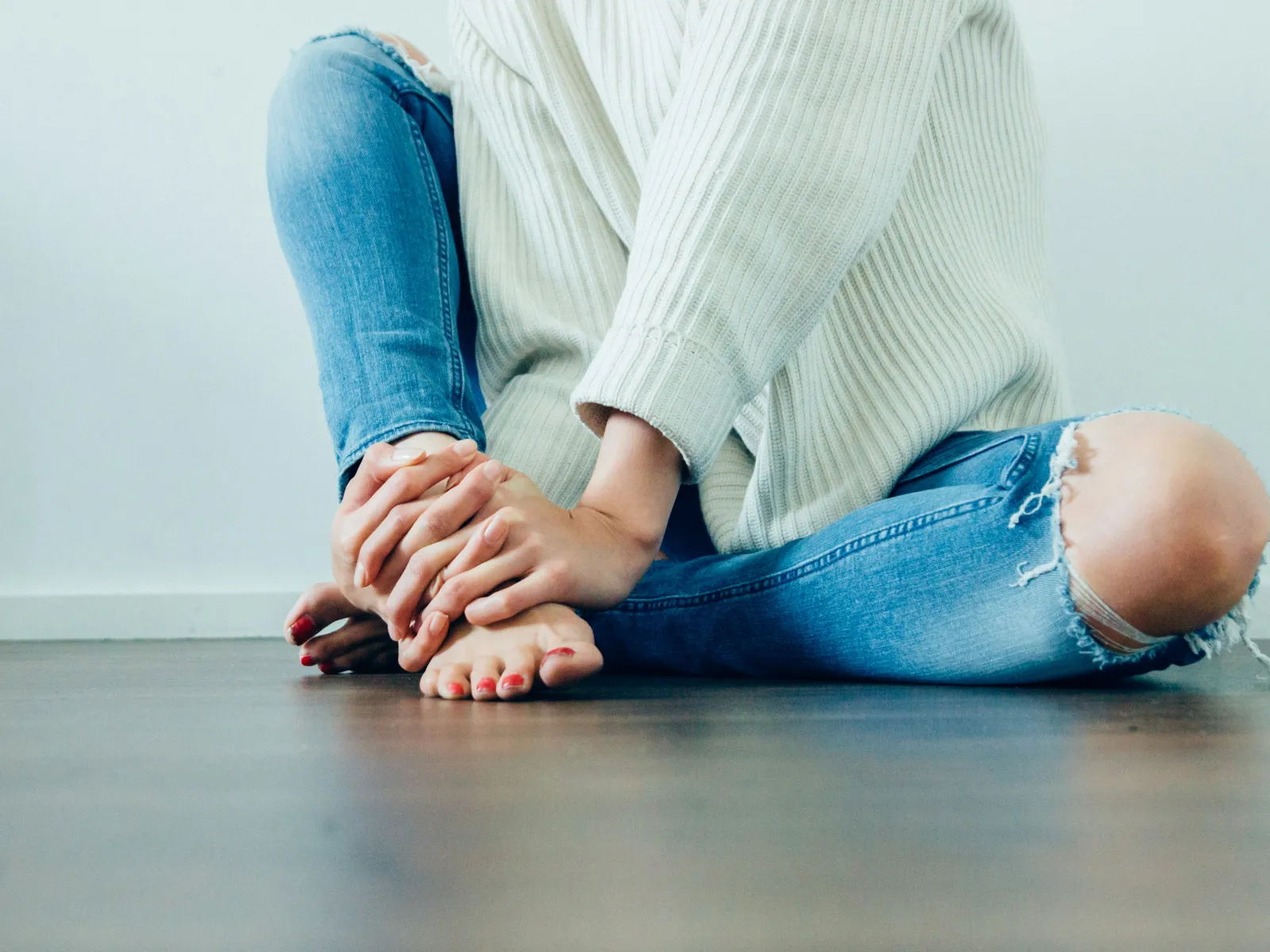 Person wearing ripped blue jeans and a white sweater sitting barefoot on wooden floor holding their foot.