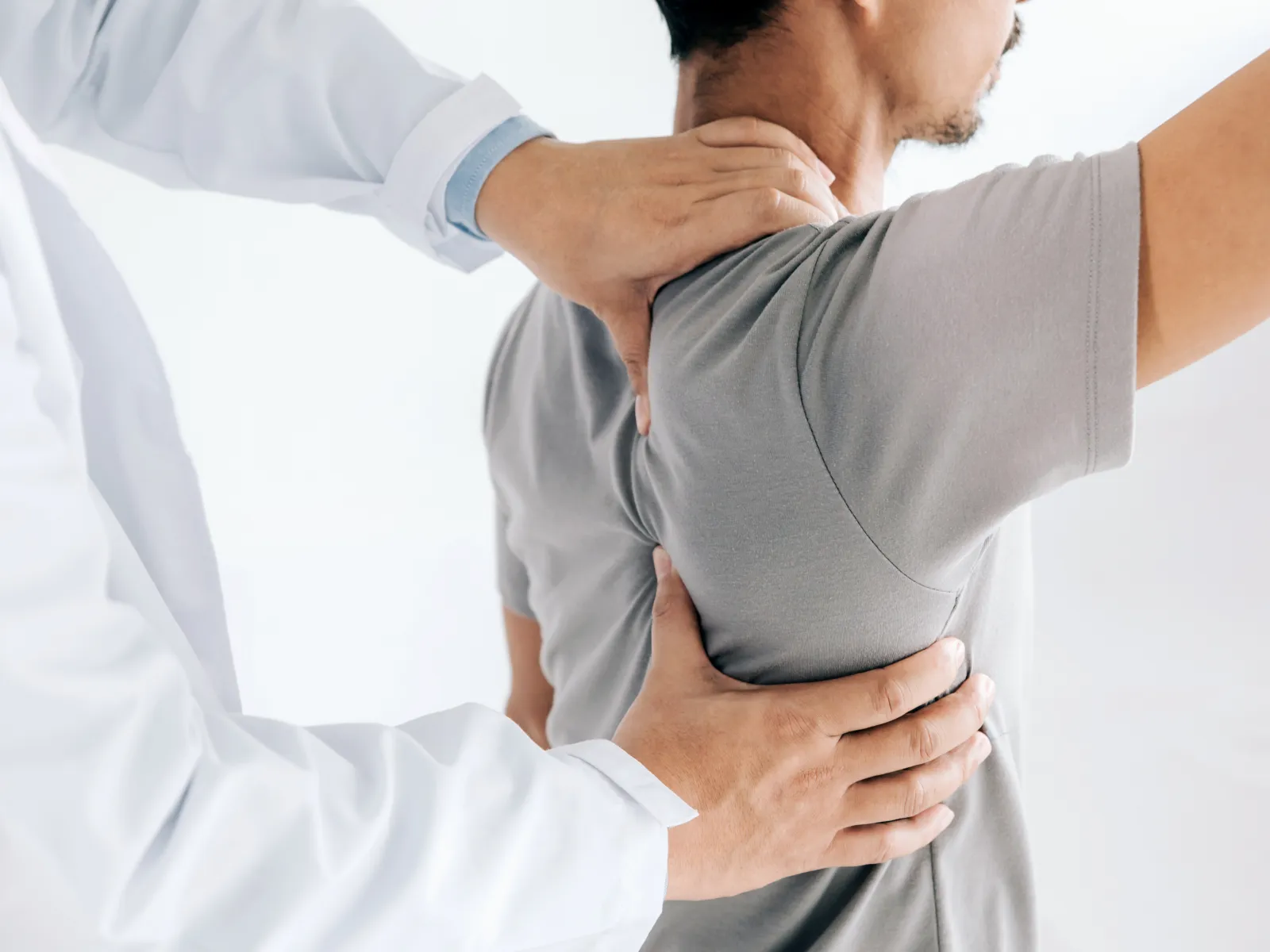 Doctor examining male patient's back and shoulder for pain or mobility issues in a clinical setting.