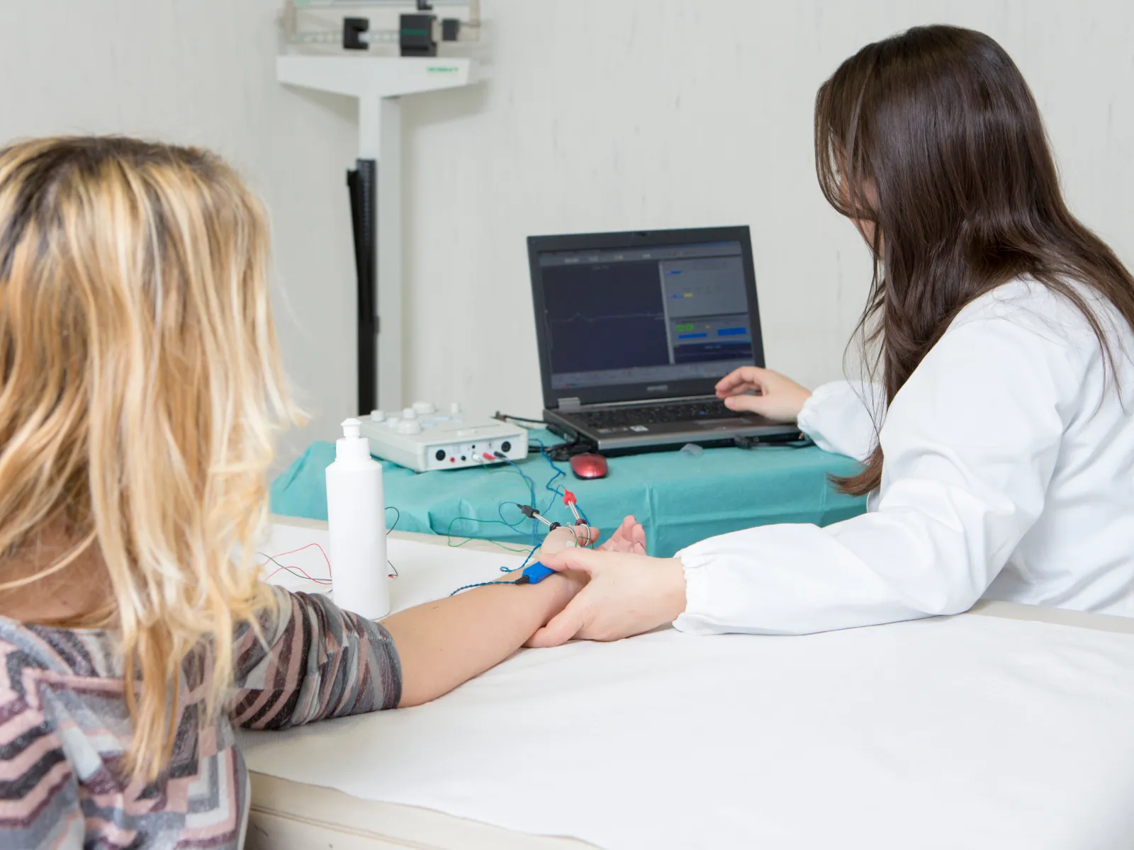 Medical professional performing electromyography test on patient using electrodes and laptop for nerve activity analysis.