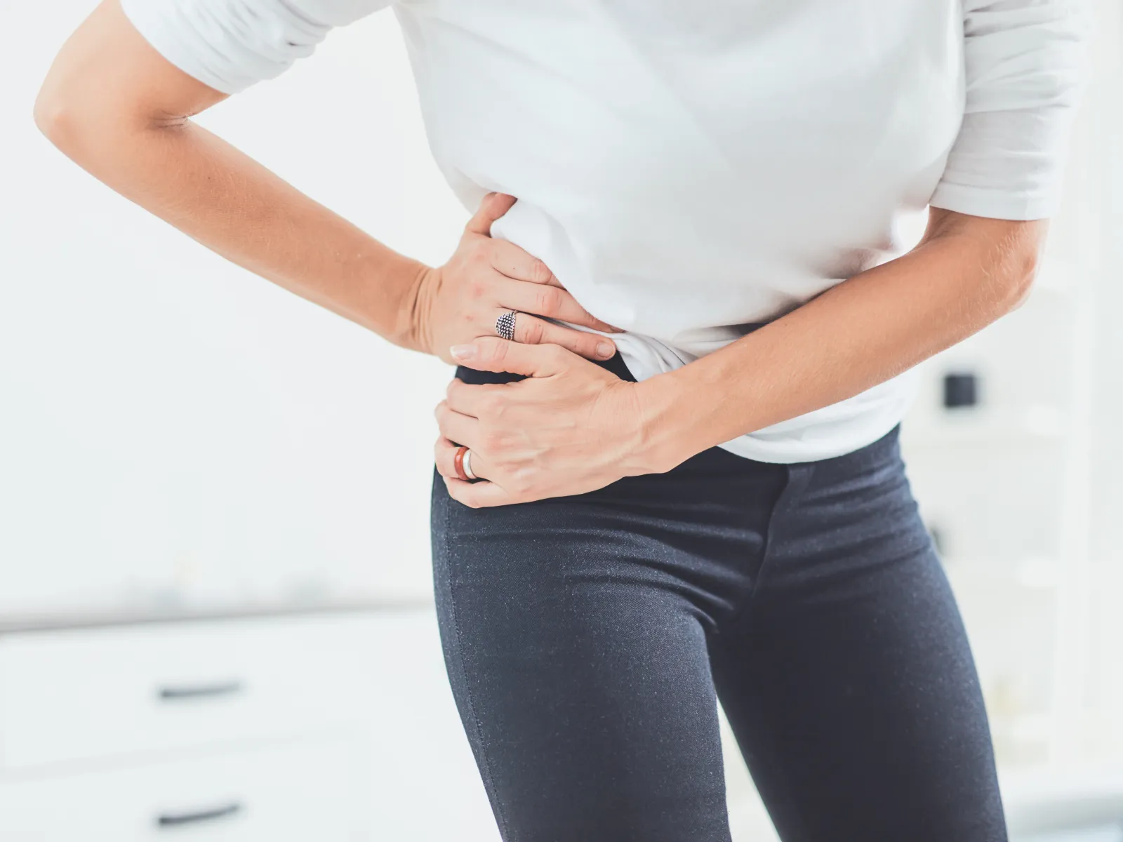 Person wearing white shirt and dark pants clutching painful hip area indoors.