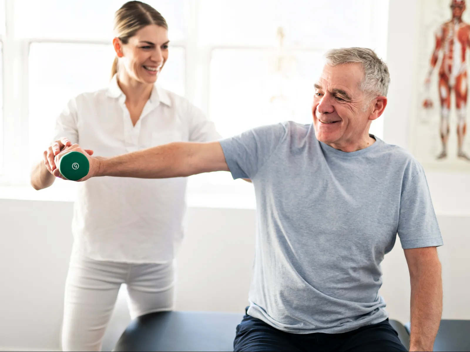 Senior man exercising with a dumbbell assisted by a smiling female physiotherapist in a bright clinic.