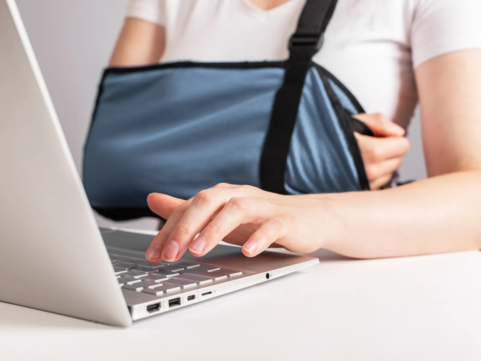 Person with arm in sling typing on a laptop at a white table, showing injury and work activity.