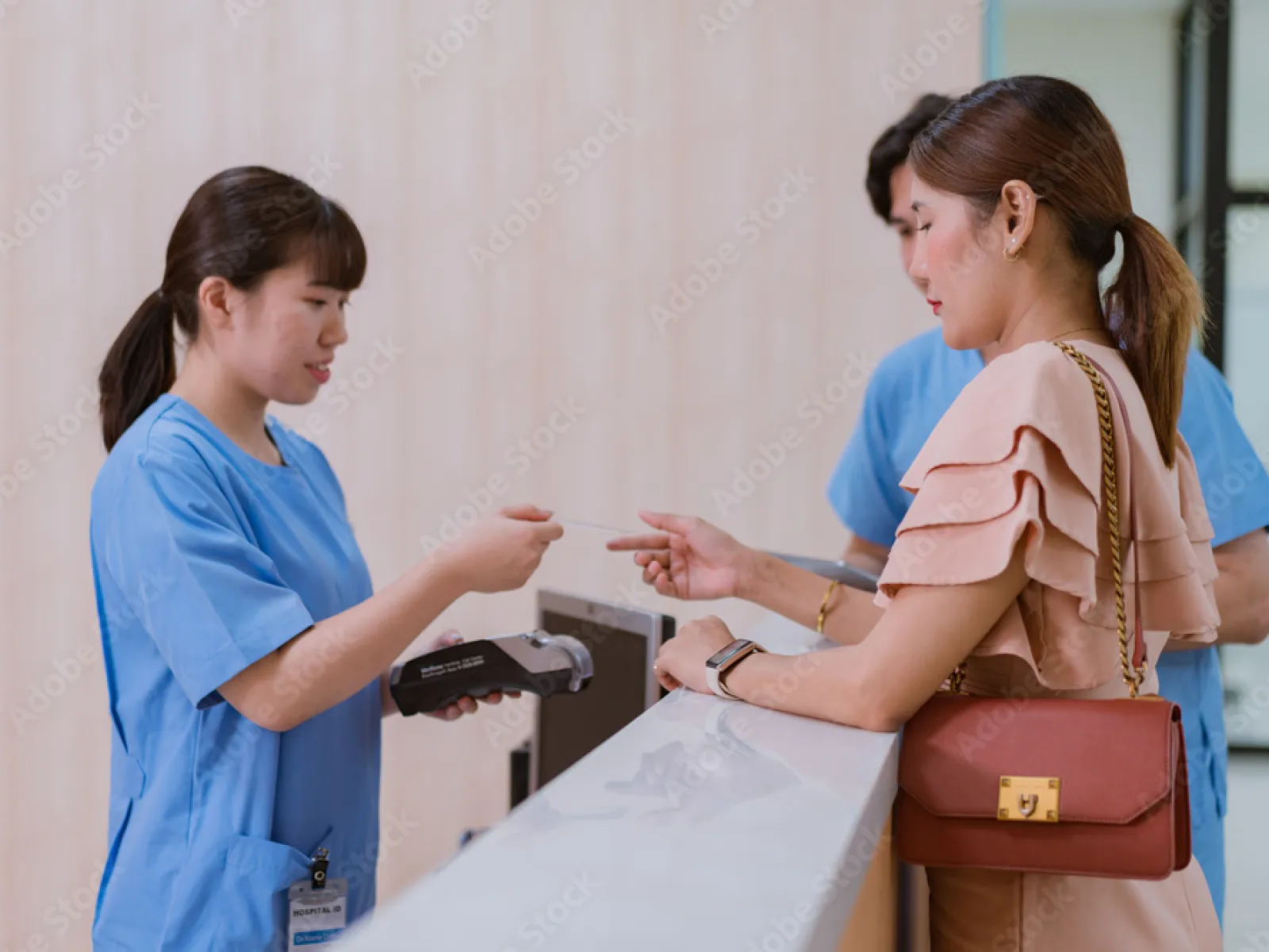 Receptionist in blue uniform handing a card to a woman with a red bag at a medical facility counter.