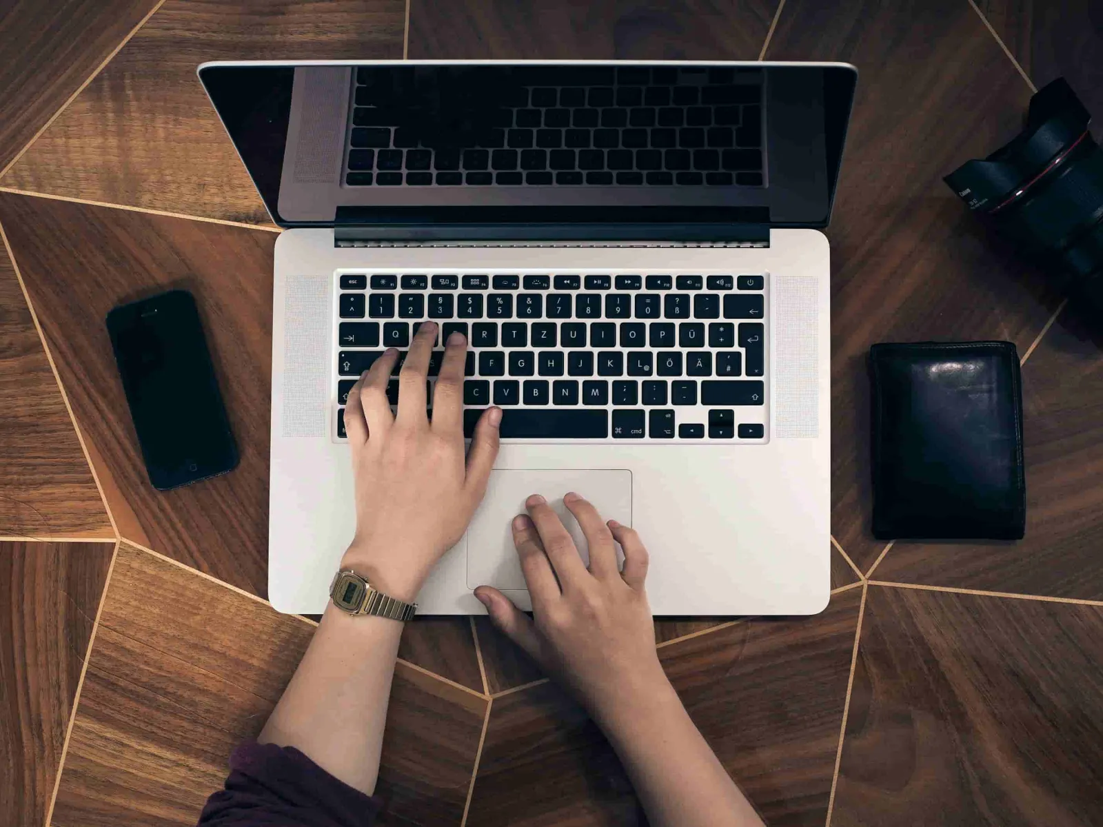 Person typing on a laptop with a smartphone, camera lens, and wallet on a wooden table.