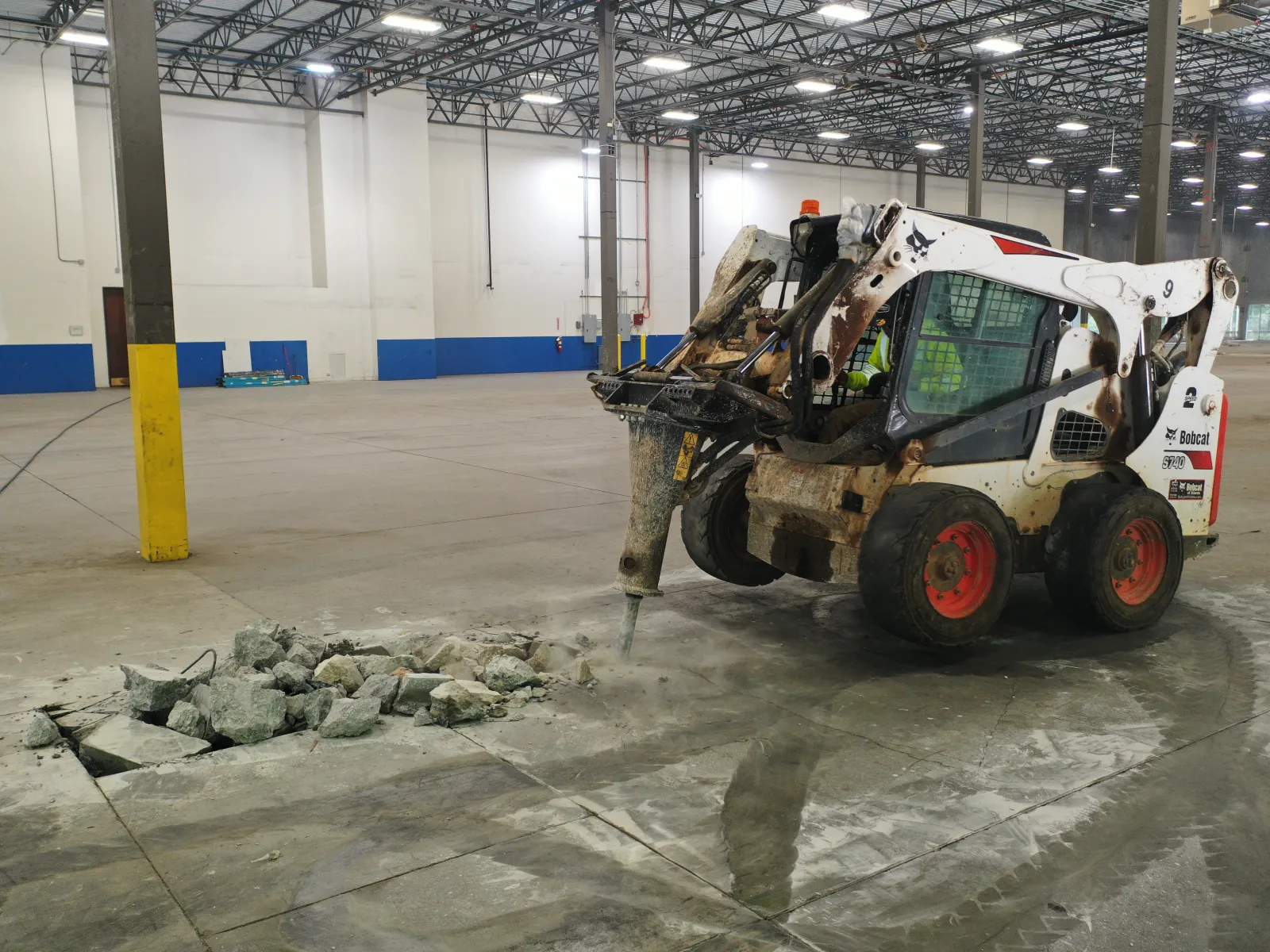 Skid steer loader breaking concrete floor inside large industrial warehouse with hydraulic breaker attachment