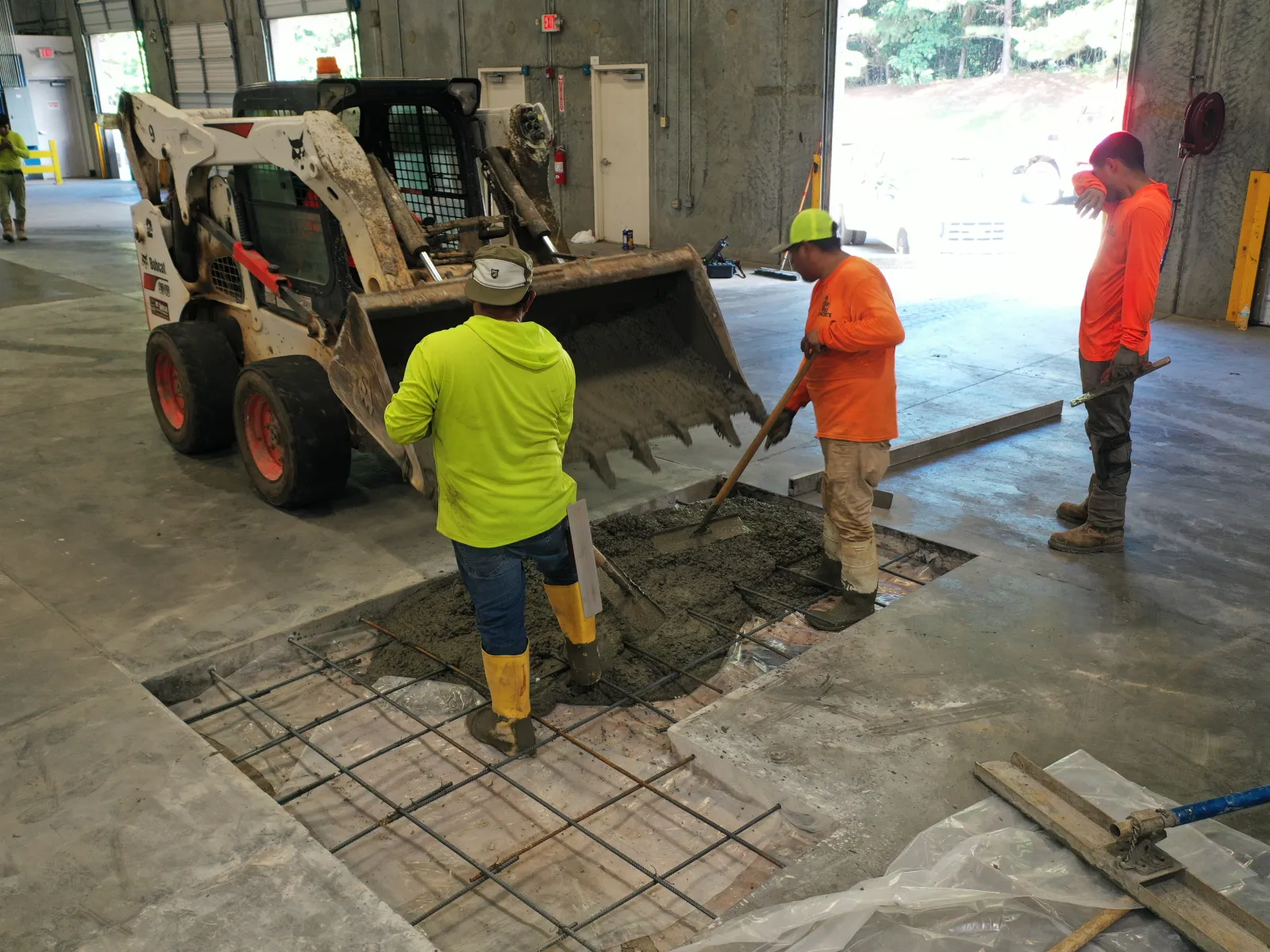 Construction workers pouring and leveling concrete inside an indoor industrial space with a skid steer loader.