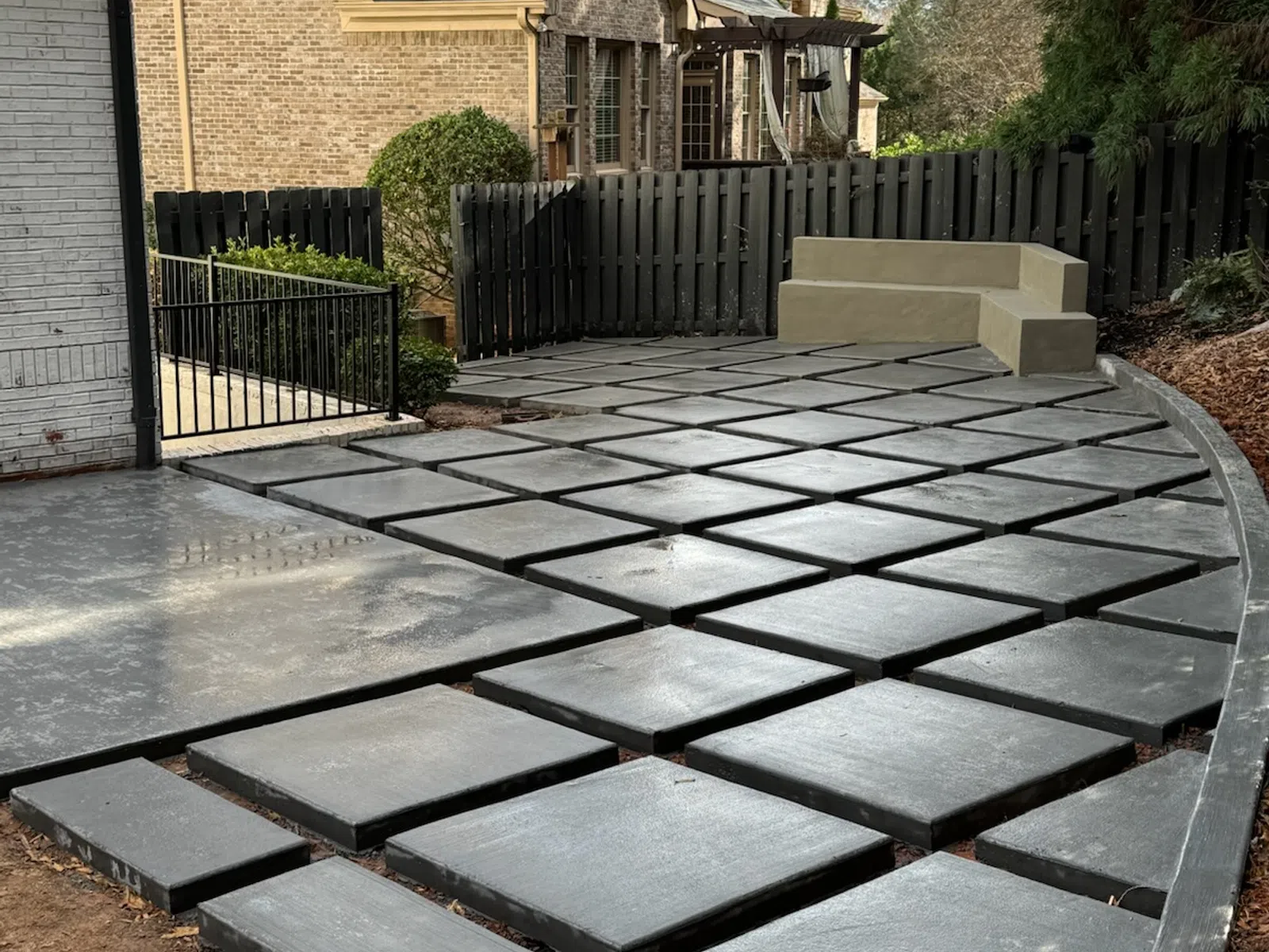 Modern backyard patio with geometric concrete pavers and built-in corner seating surrounded by a black fence.