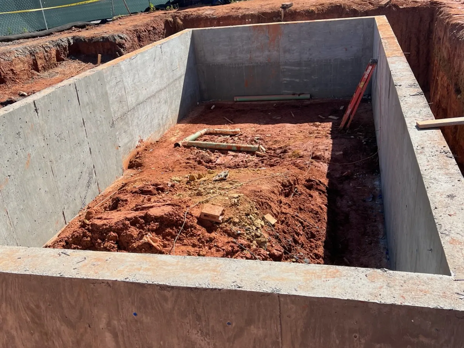 Concrete foundation pit with exposed soil and pipes at construction site surrounded by fencing and trees.