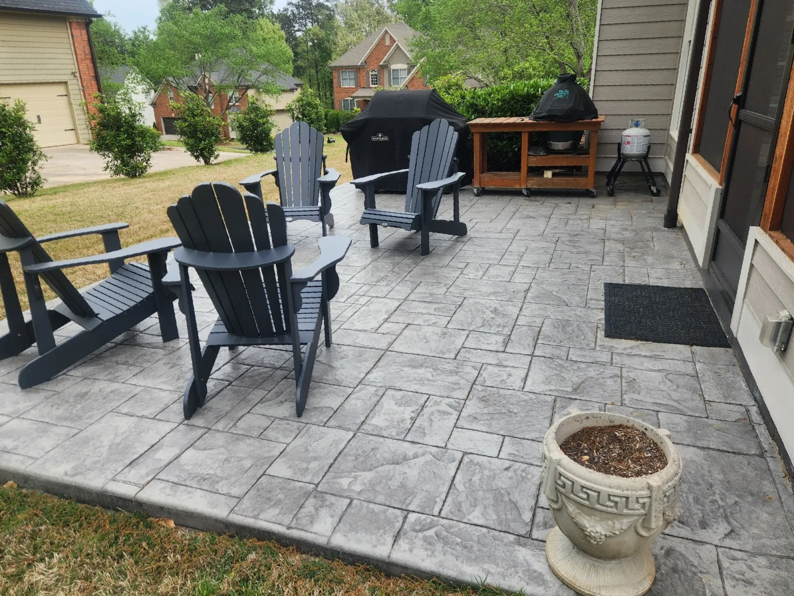 Relaxing outdoor patio with gray stamped concrete, Adirondack chairs, and a grill in a leafy neighborhood.