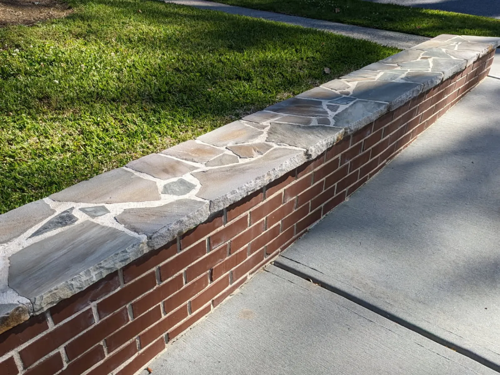 Low brick and stone wall bordering a green lawn and adjacent to a concrete sidewalk on a sunny day