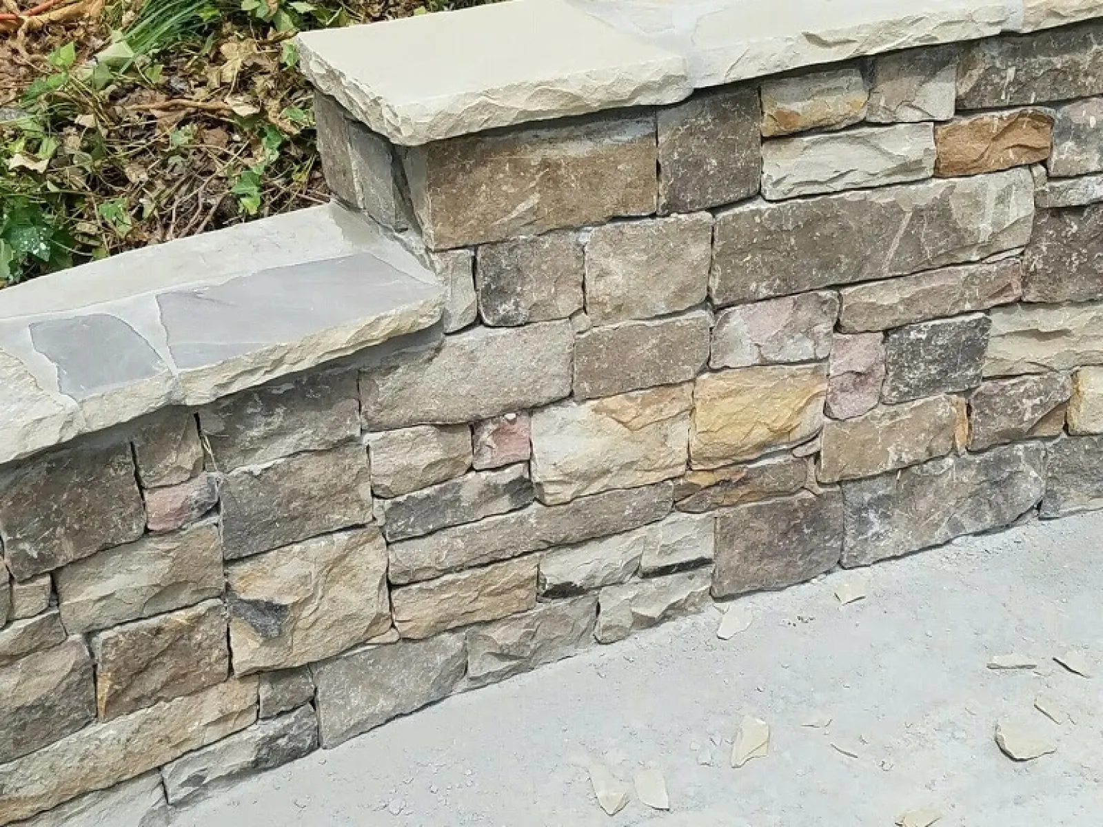 Curved stone retaining wall with natural stone veneer and concrete base next to vegetation and grass.