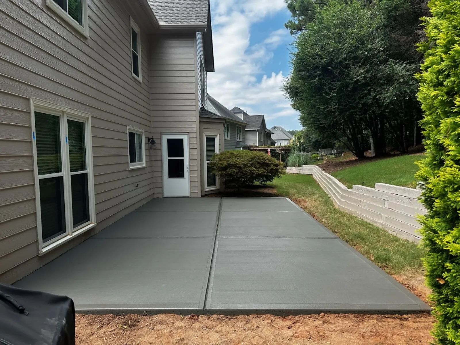 Gray concrete patio beside a house, surrounded by greenery and a clear blue sky.