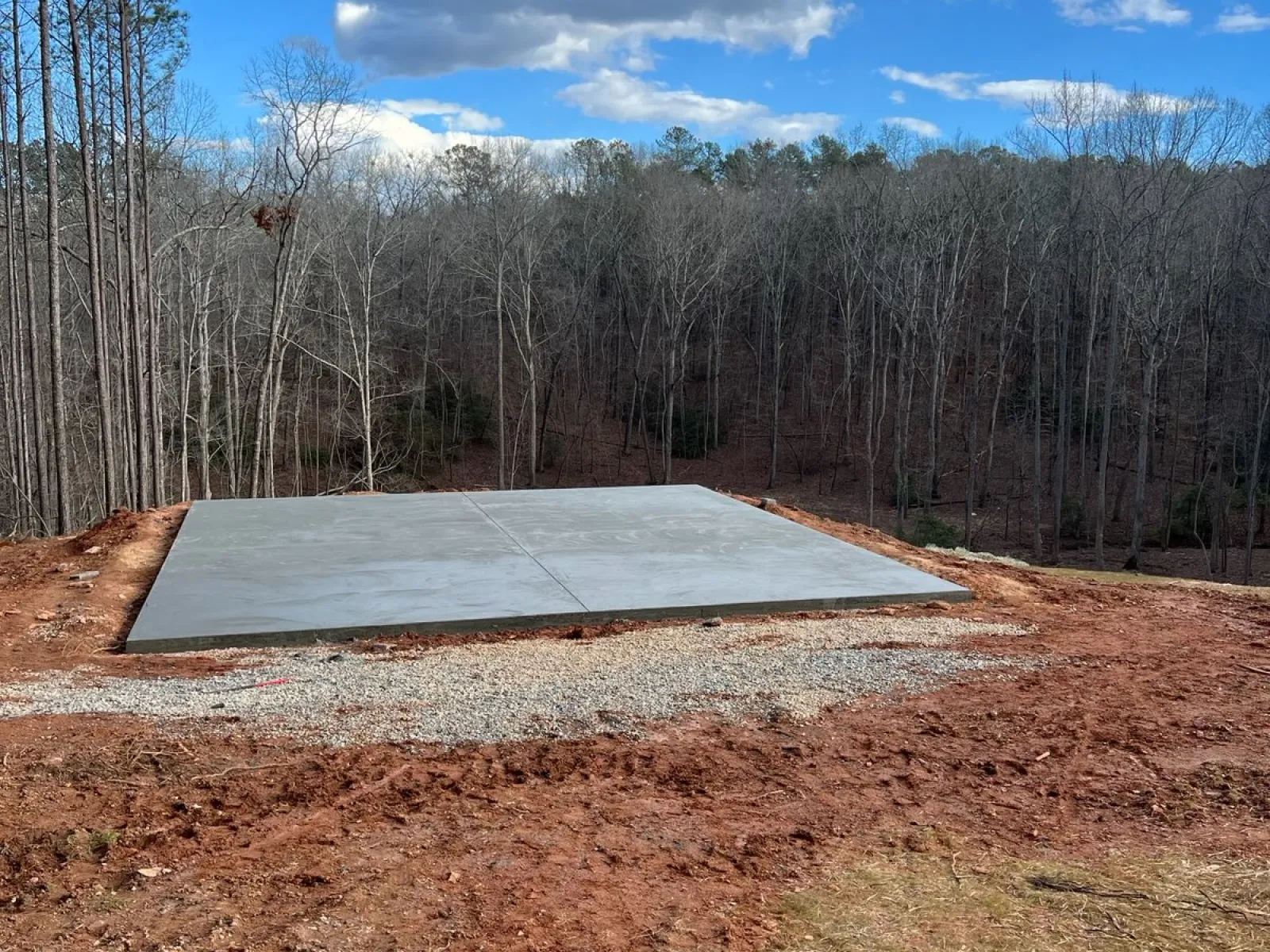 Newly poured concrete slab foundation on cleared soil with a forest and cloudy blue sky in the background