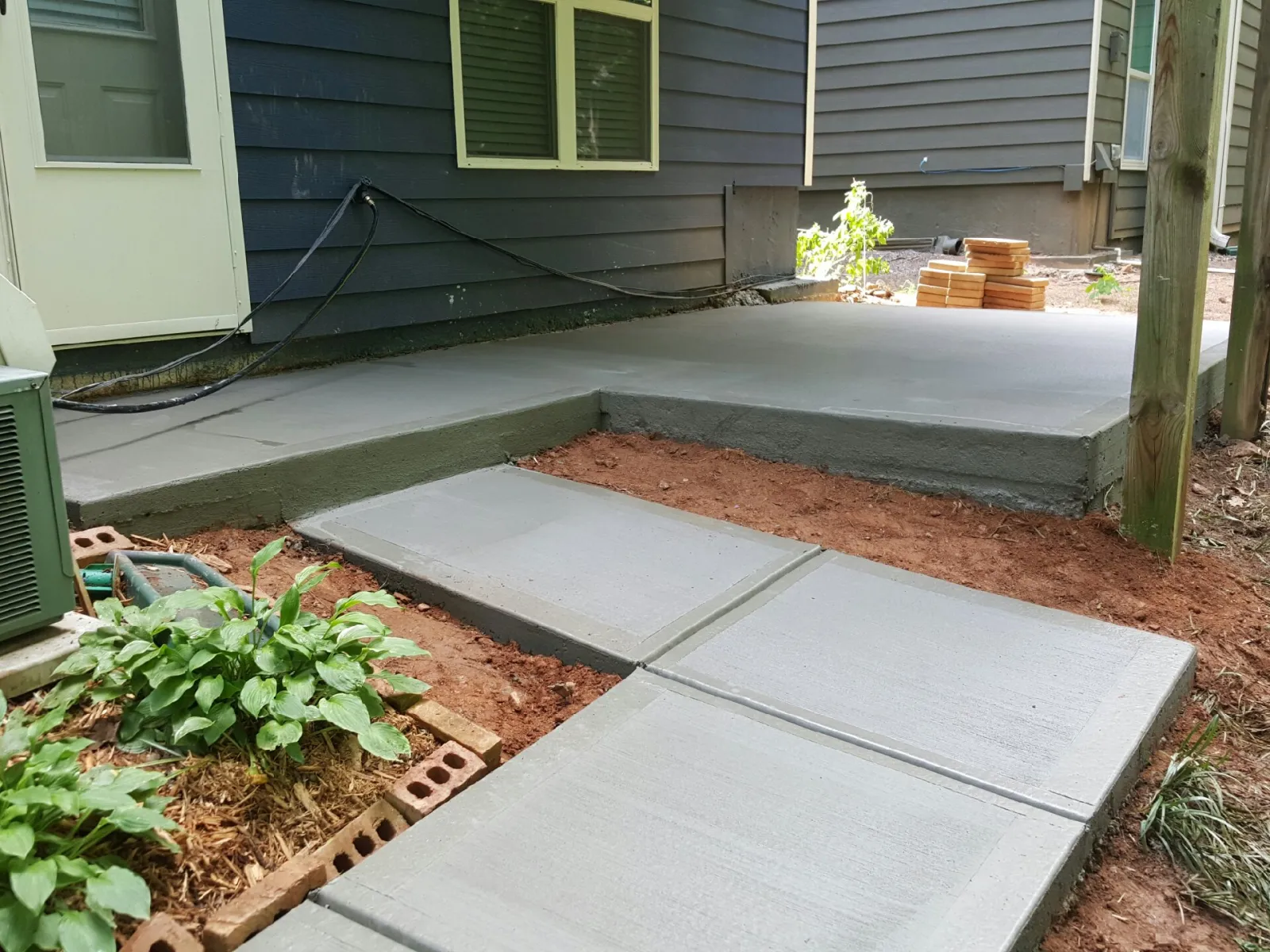 Freshly poured concrete patio and walkway beside a modern home, featuring landscaping elements.