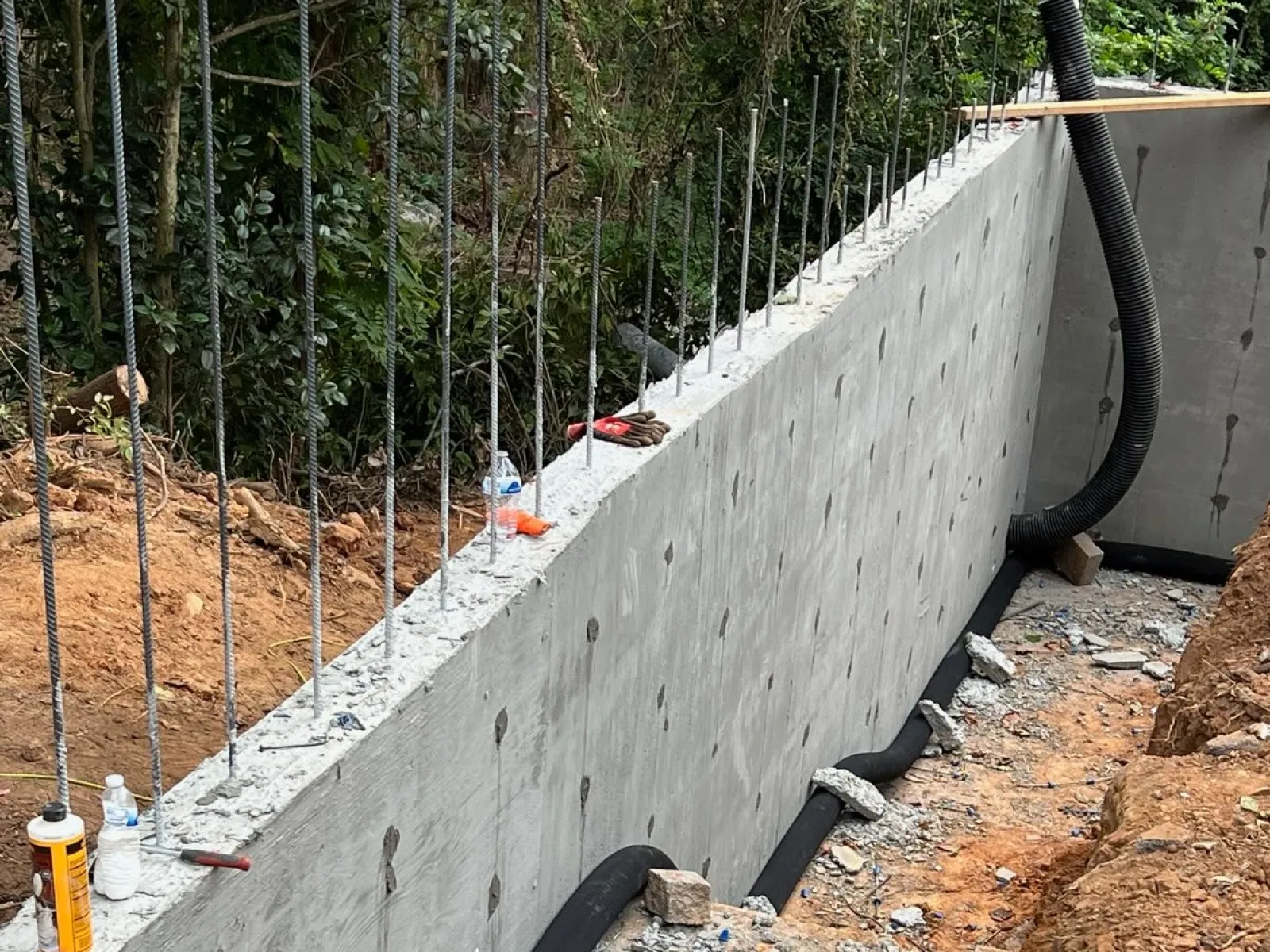 A newly constructed concrete wall with steel reinforcement bars on a construction site surrounded by dirt and tools.