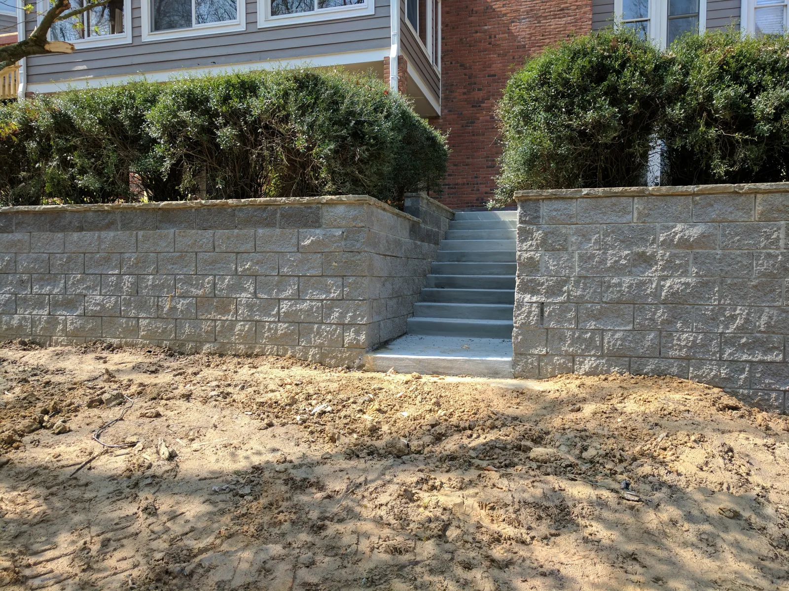 Concrete retaining walls and steps with bushes on top in front of a residential house with bare soil in foreground