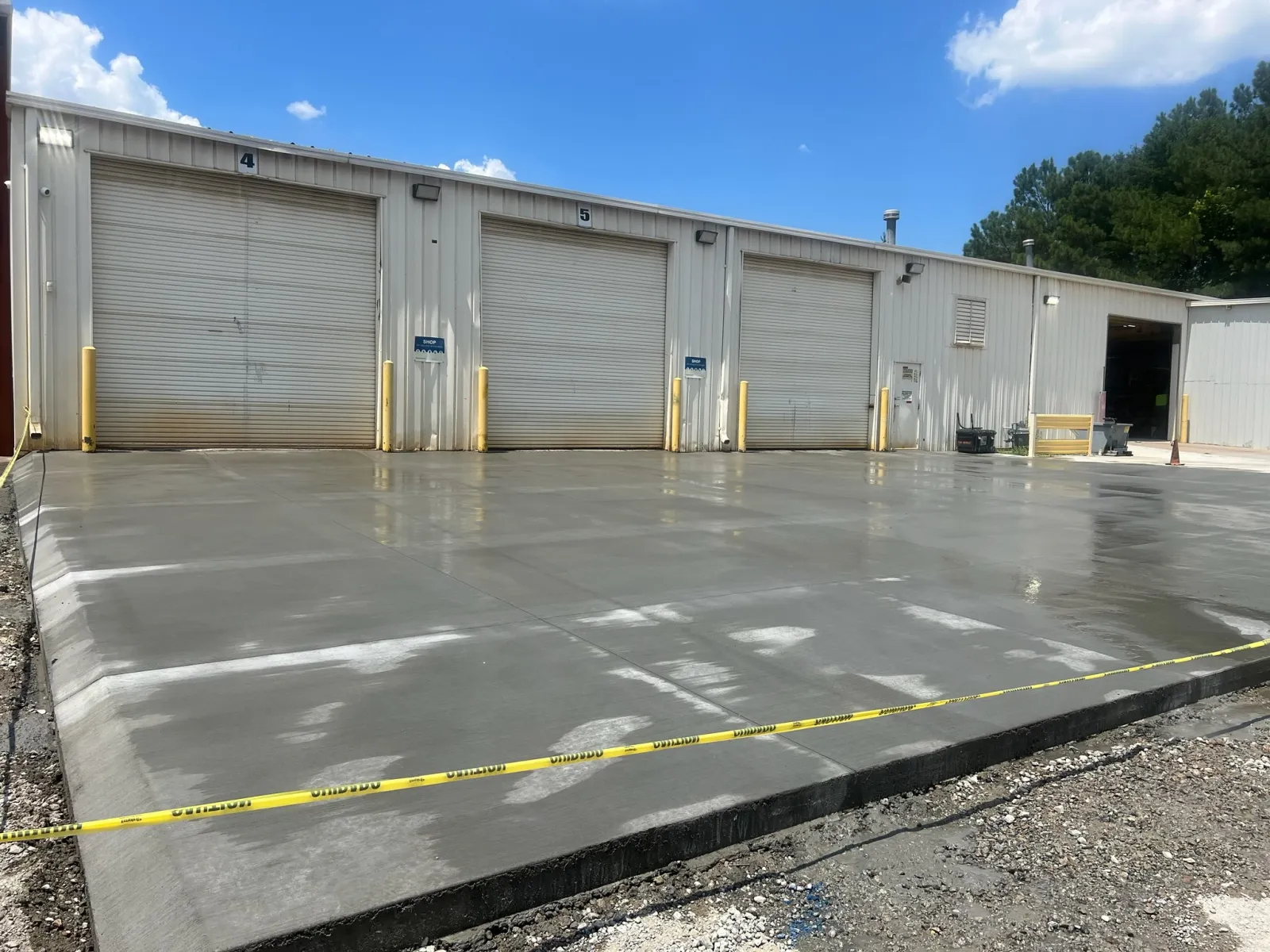 Newly poured concrete slab outside warehouse with three closed loading dock doors and caution tape.