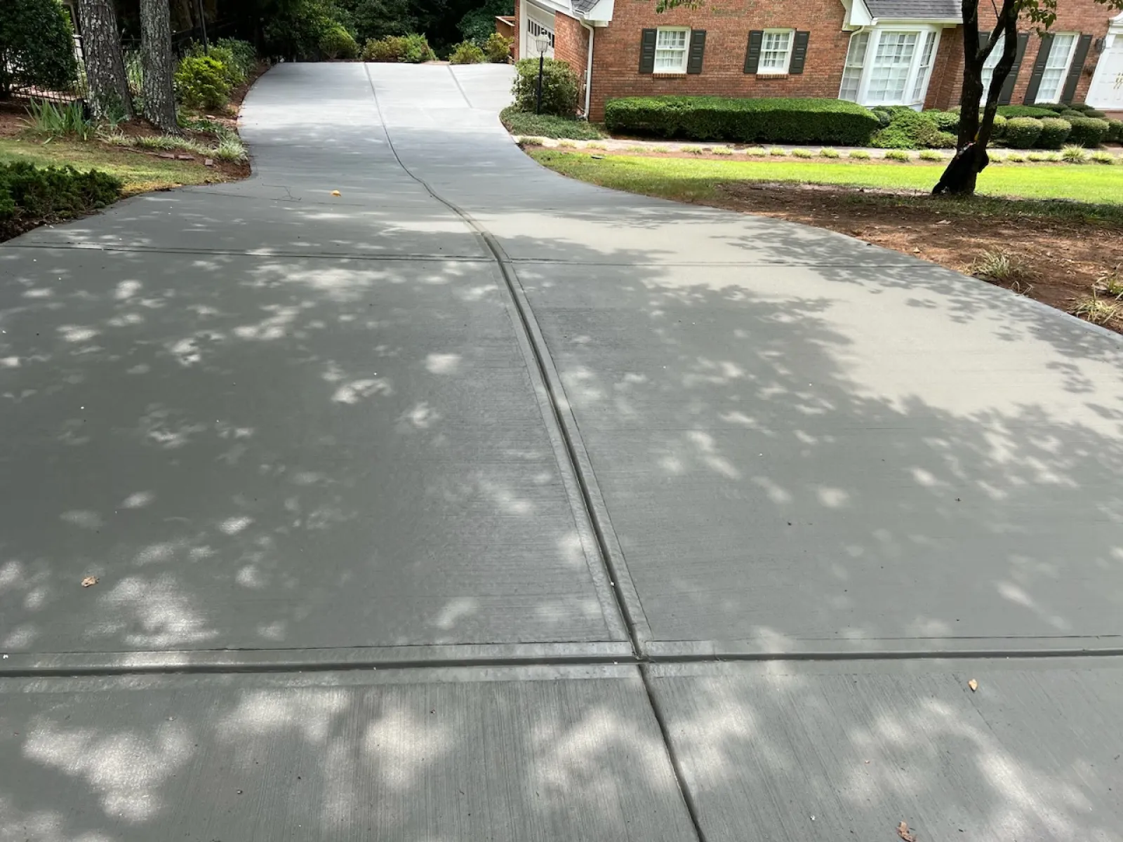 Newly poured concrete driveway leading to a brick house with surrounding greenery and tree shadows.