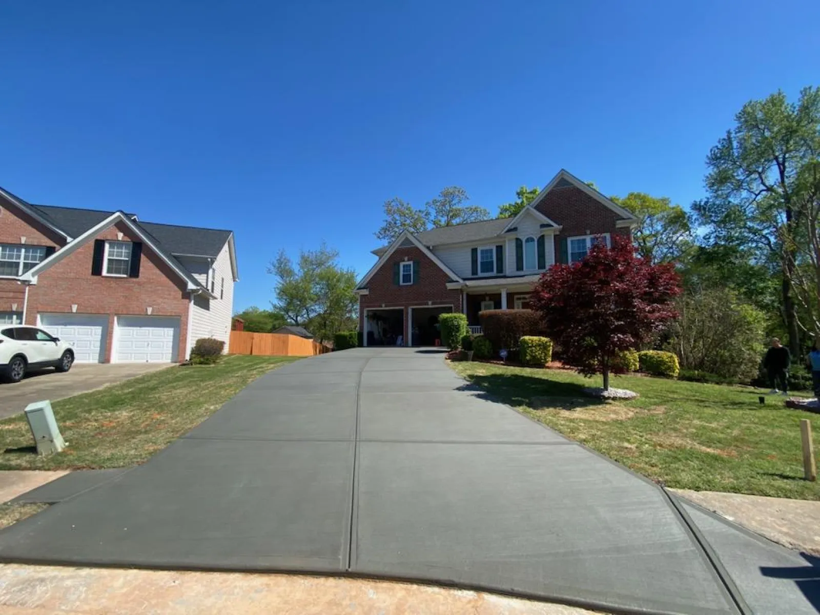 Wide concrete driveway leading to a two-story brick house with a red tree and green lawn under a clear blue sky
