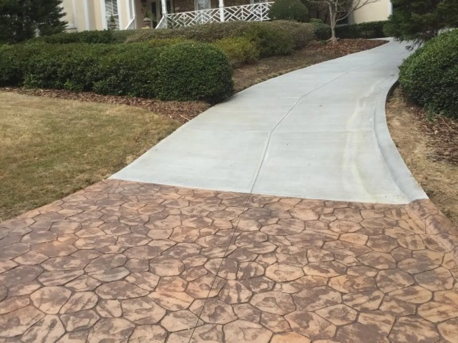 Concrete driveway with stamped stone design meets a smooth gray walkway in a landscaped setting.