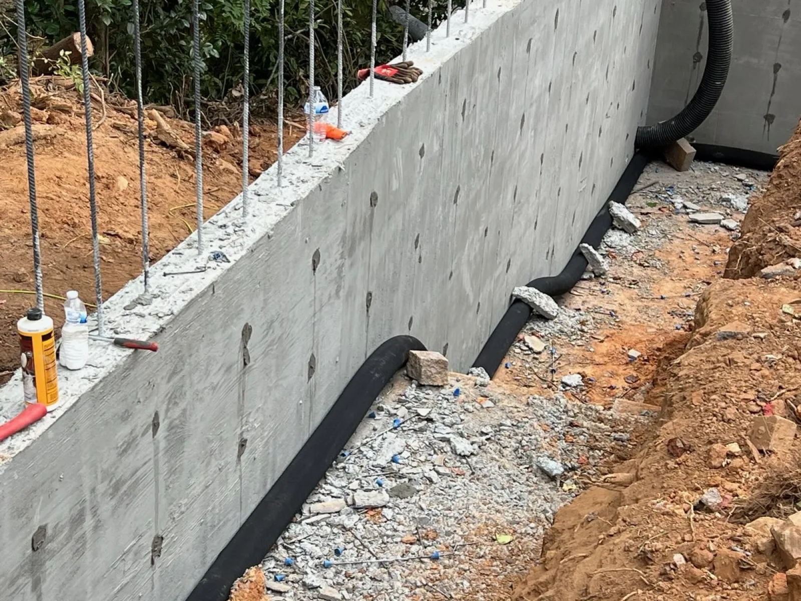 Concrete foundation wall with vertical rebar rods and drainage pipes installed in a gravel trench beside earth.