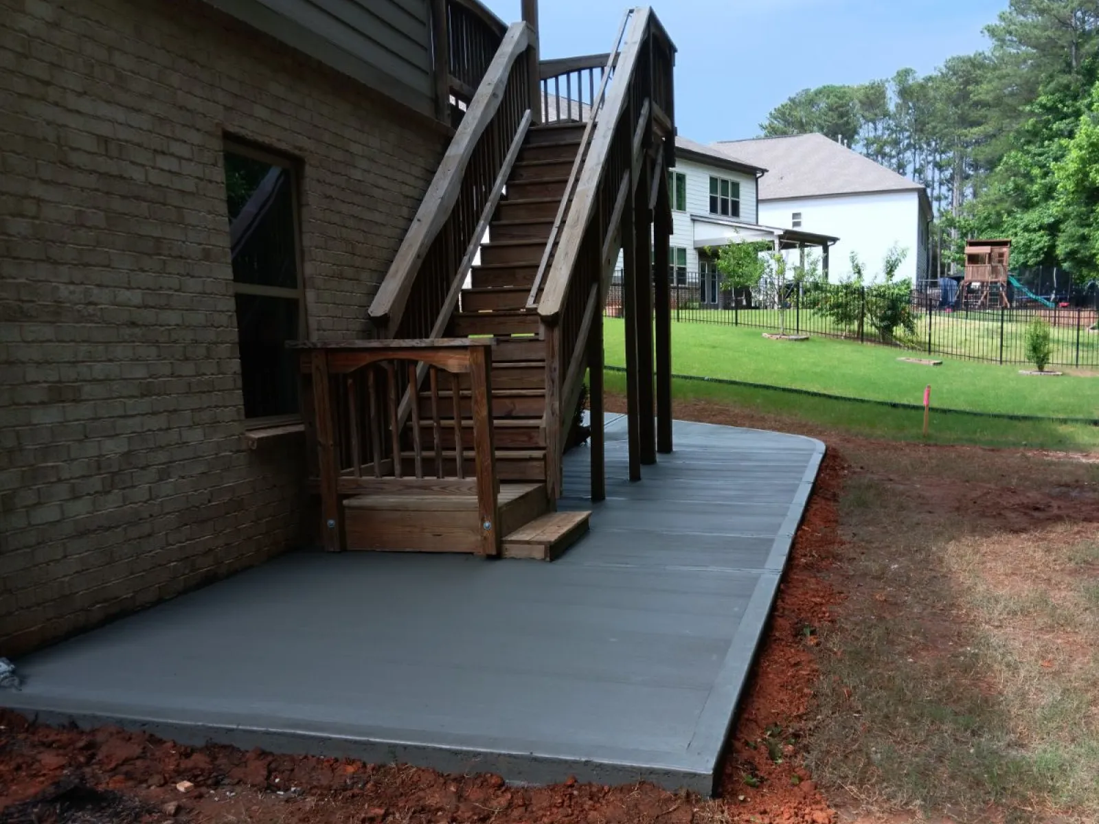 New concrete patio slab next to wooden staircase attached to a brick house in a suburban backyard.